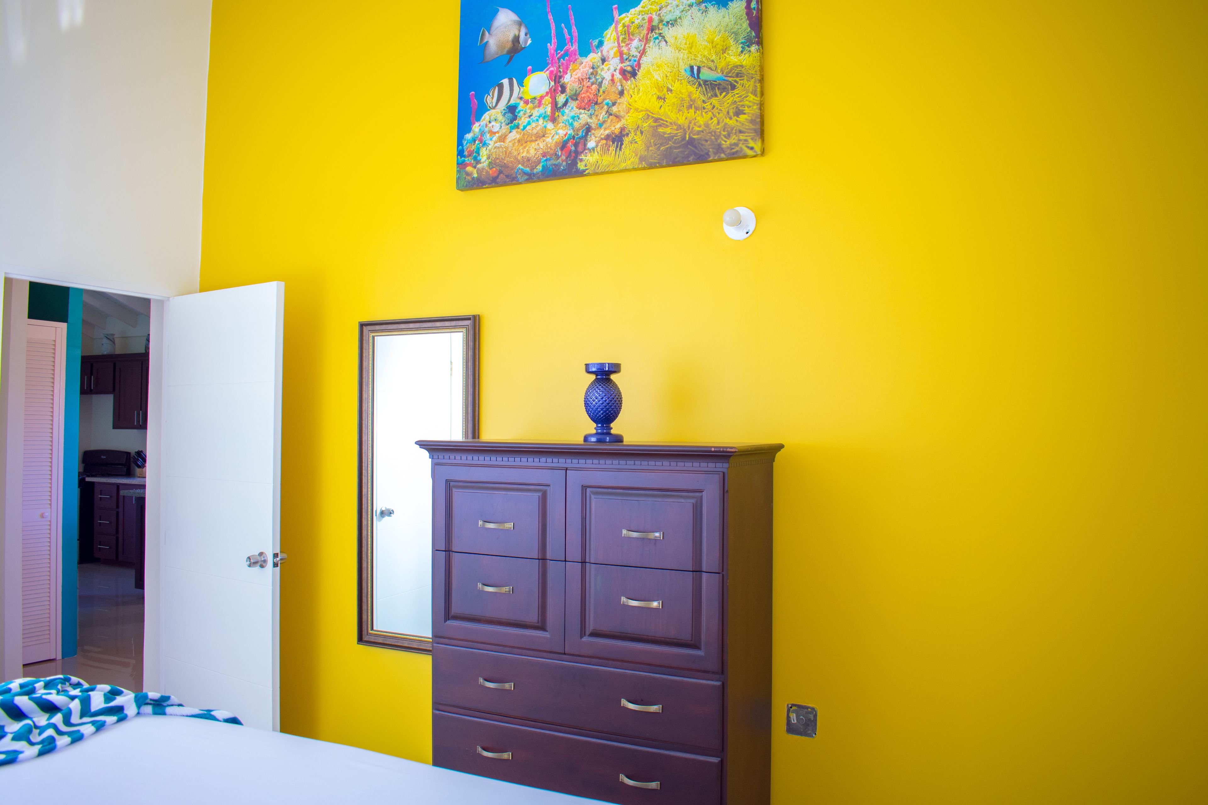 Una habitación con cama, cómoda y un cuadro de peces en una pared amarilla.
