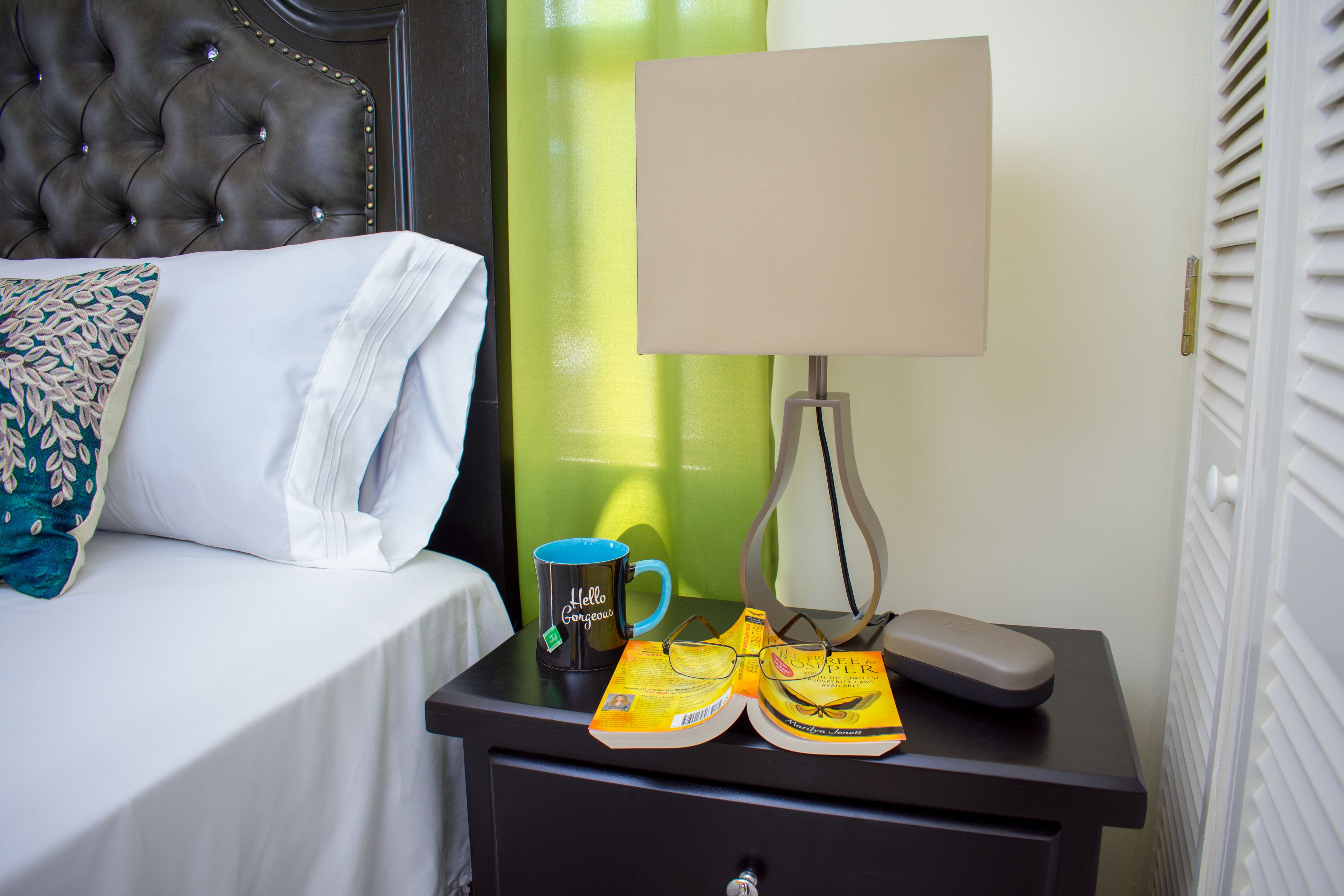 Una cama bien hecha con una colcha blanca y una almohada decorativa. Junto a la cama, una mesita de noche con una lámpara, una taza y un libro.