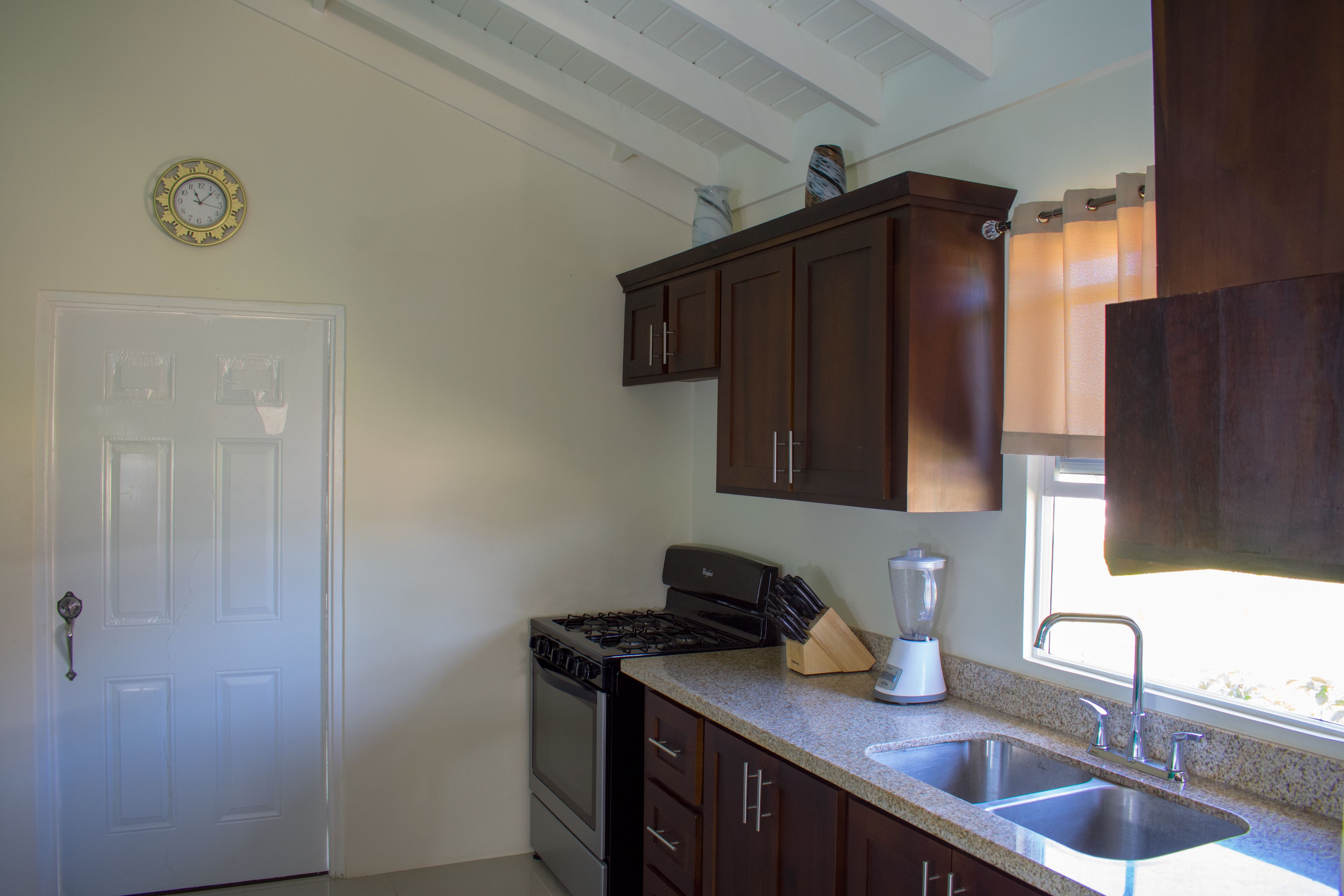 Una cocina con armarios de madera oscura, una puerta blanca, una estufa, un fregadero y un reloj en la pared.