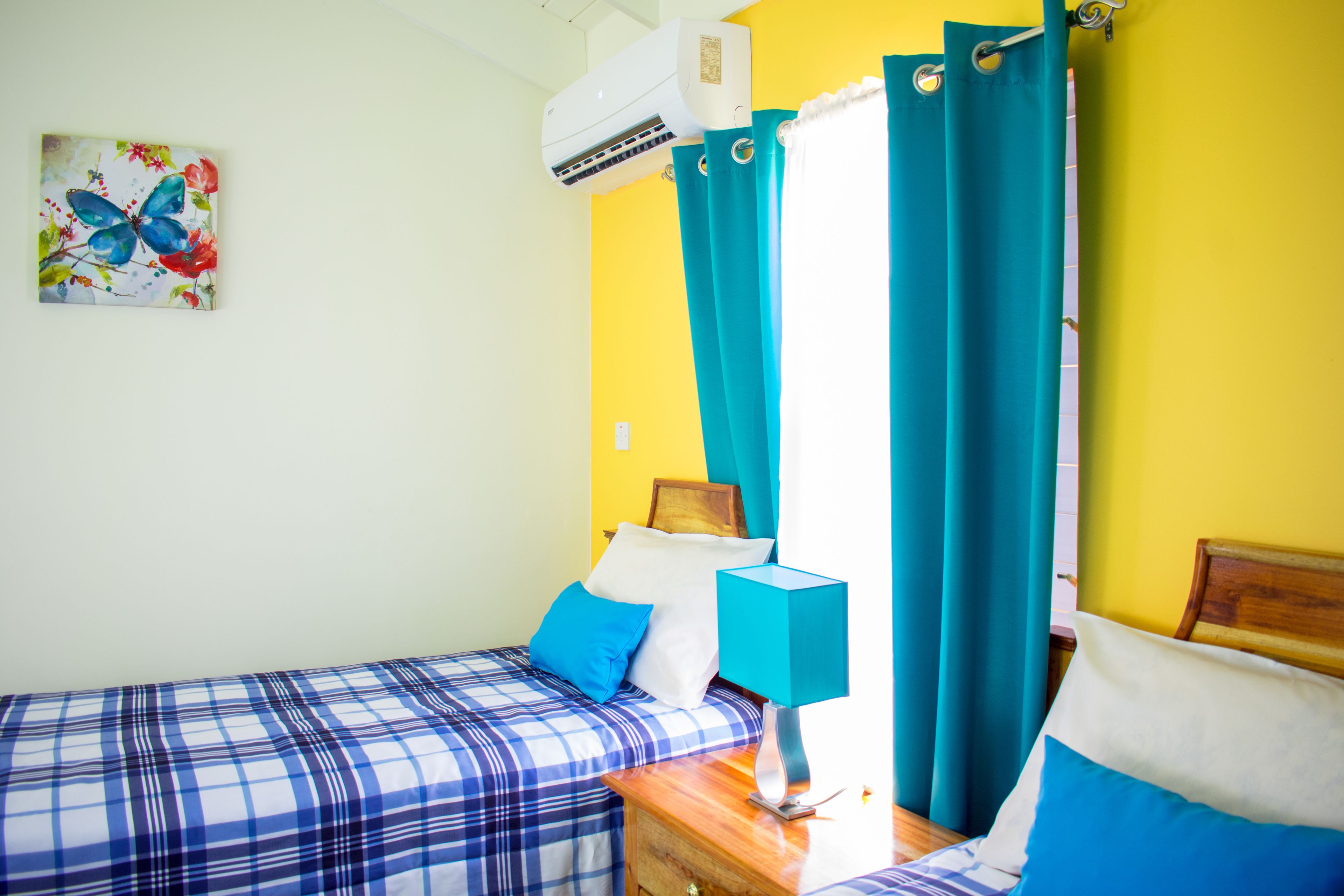 A room with two beds, a nightstand with a lamp, a wall-mounted air conditioner, and a painting of a butterfly.