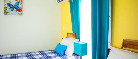 A room with two beds, a nightstand with a lamp, a wall-mounted air conditioner, and a painting of a butterfly.