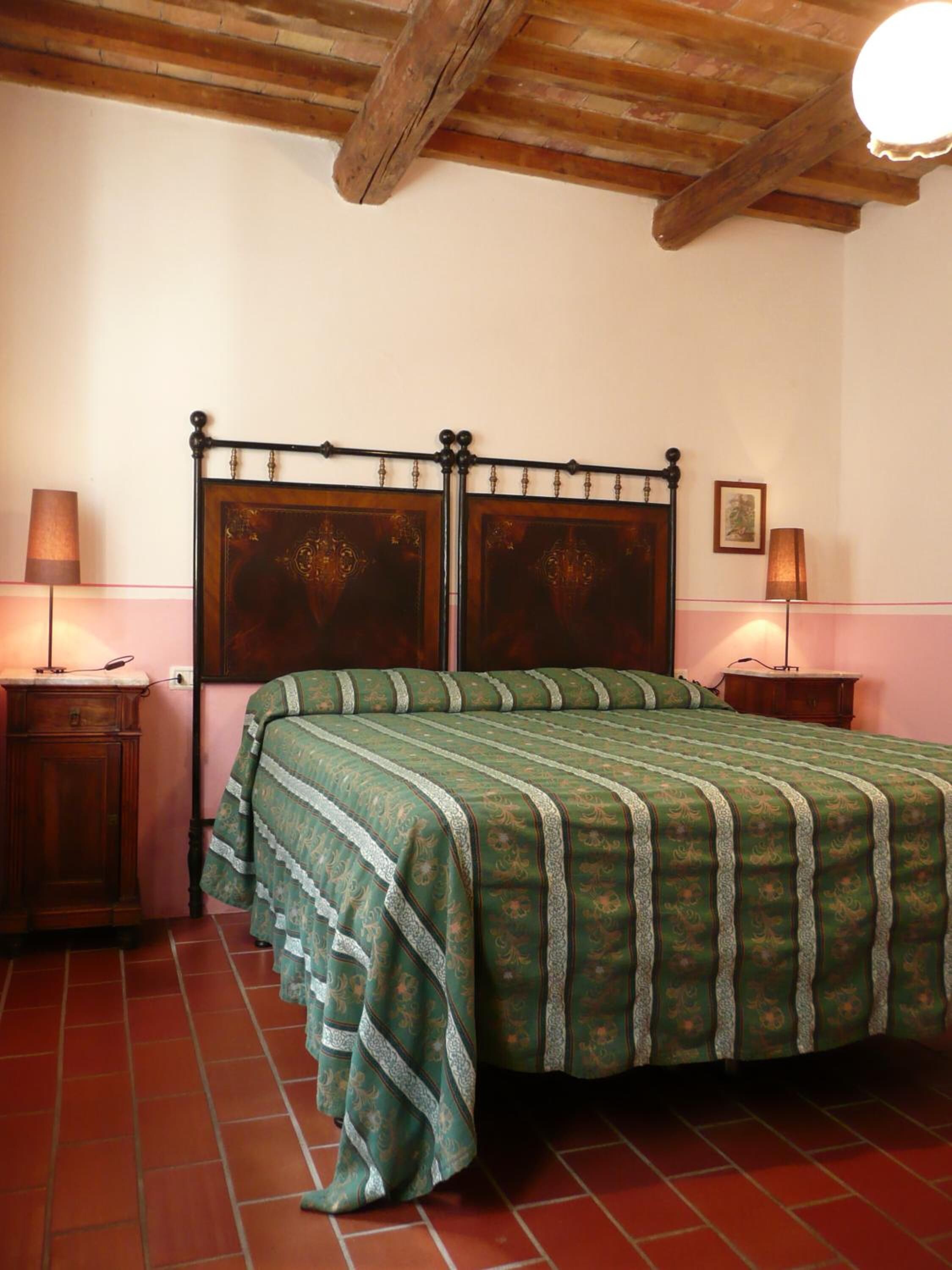 A bedroom with a wooden ceiling, a bed with a green patterned bedspread, two bedside tables with lamps, and a framed picture on the wall.