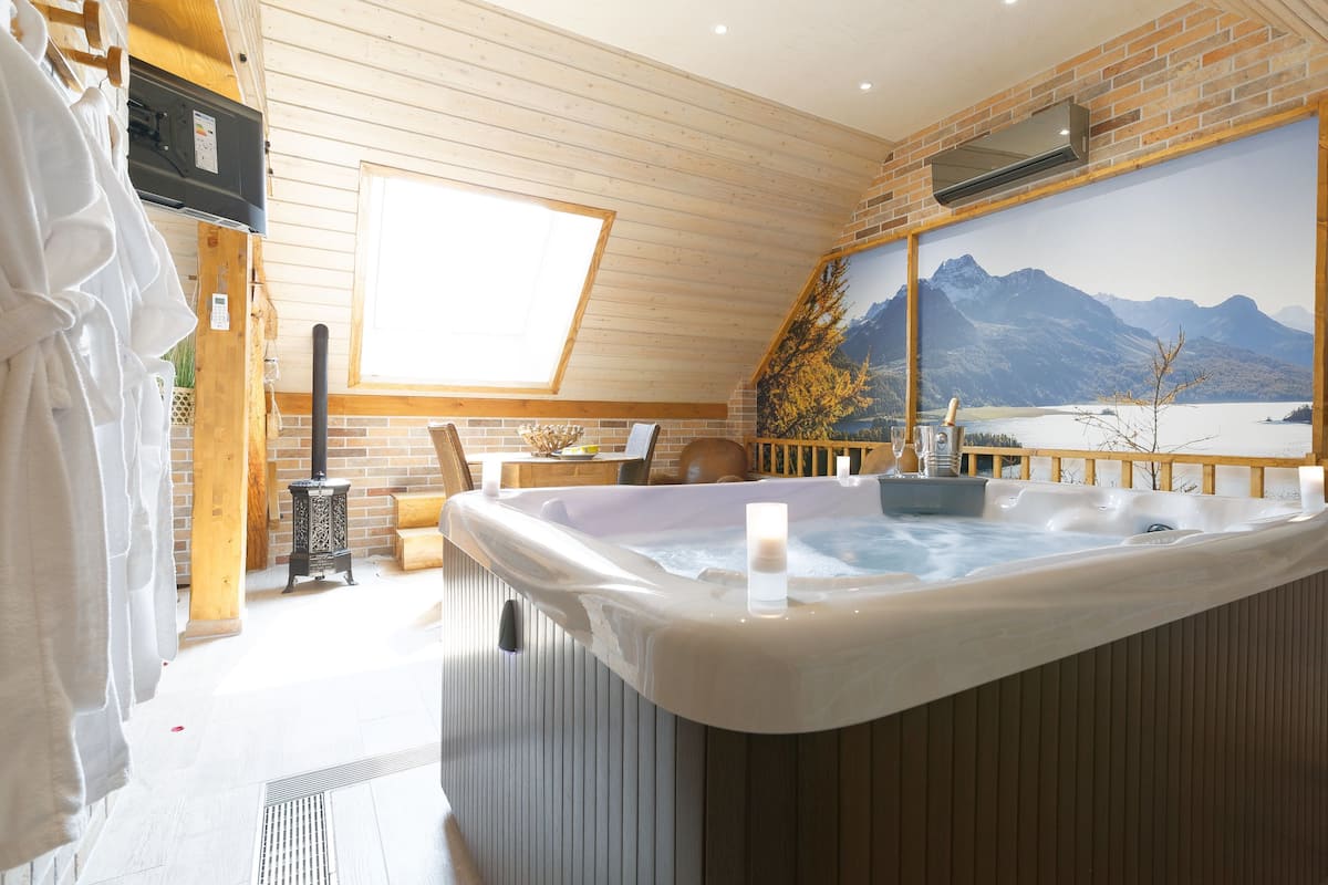 A spa room with a hot tub, wooden walls, a large window, and a mountain view painting.