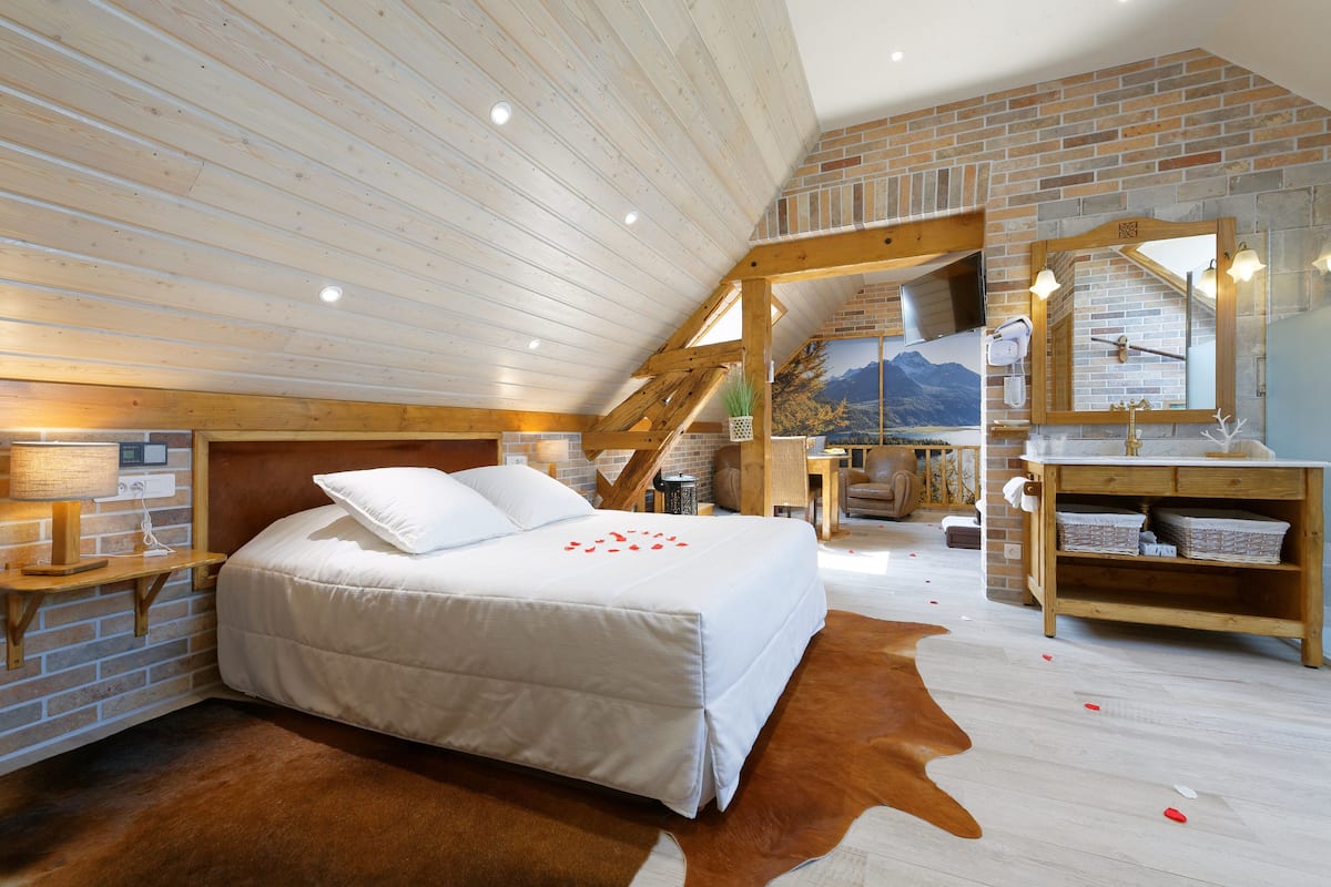 A bedroom with a wooden ceiling, brick walls, a bed with white bedding, a wooden bench, a mirror, and a bathroom with a wooden vanity.