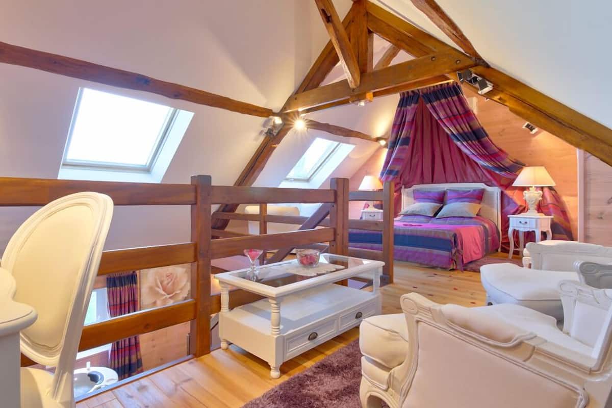 A cozy attic bedroom with a sloped ceiling, wooden beams, a bed with colorful bedding, a white sofa, a small table, and a window with curtains.