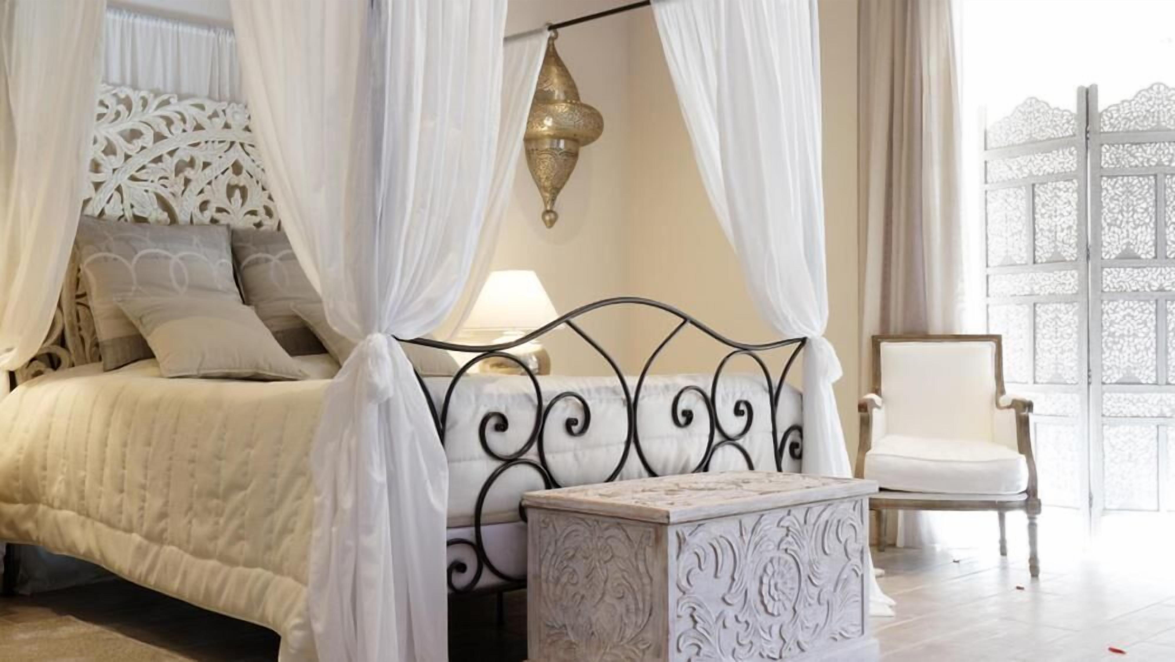 A bedroom with a four-poster bed, a nightstand, a chair, and a decorative wall piece.