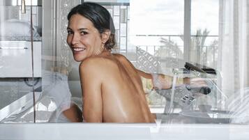 A person in a bathtub with a showerhead, a framed picture on the wall, and a window with a view.