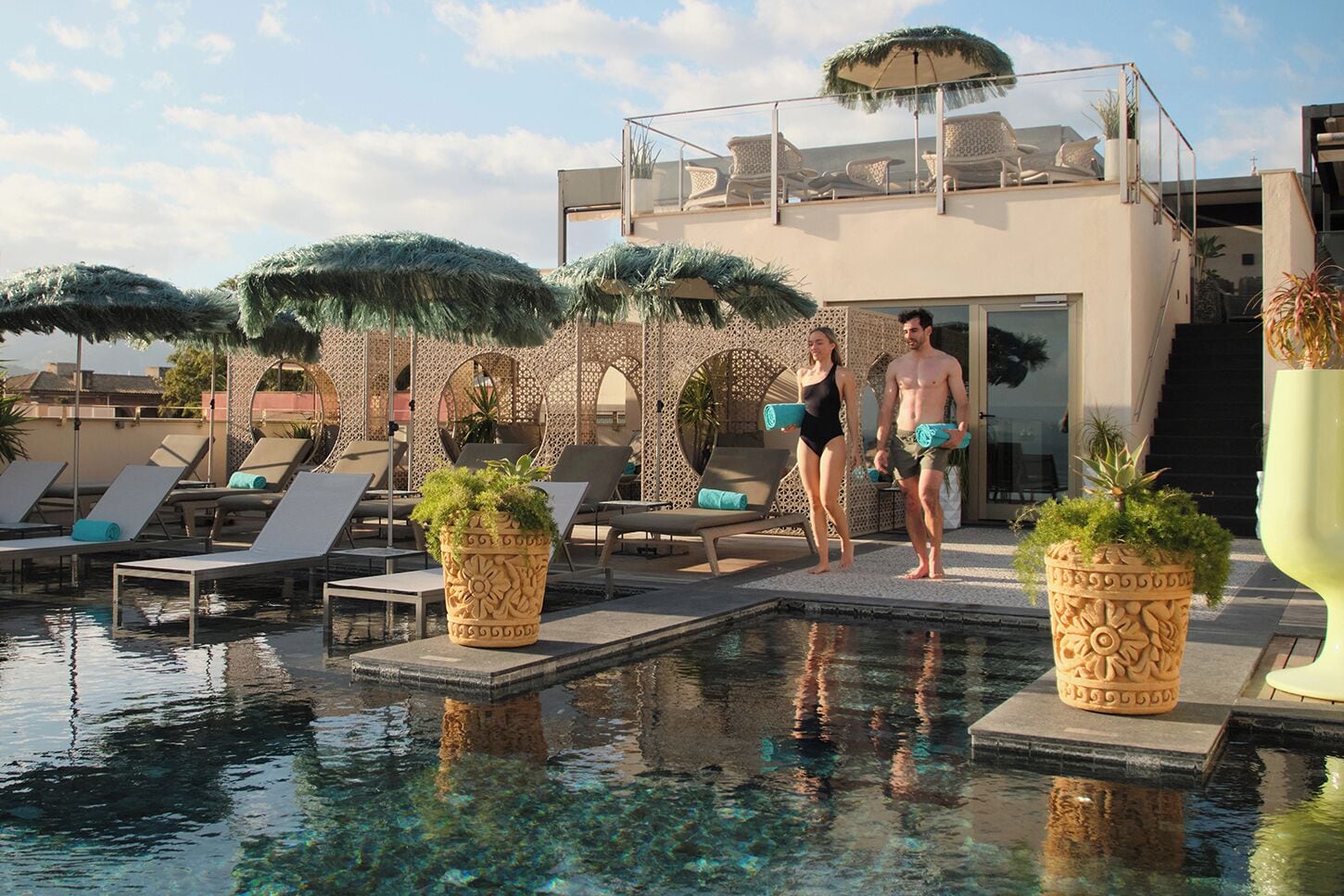 Zona de piscina con sillones reclinables, una pareja caminando y macetas decorativas.
