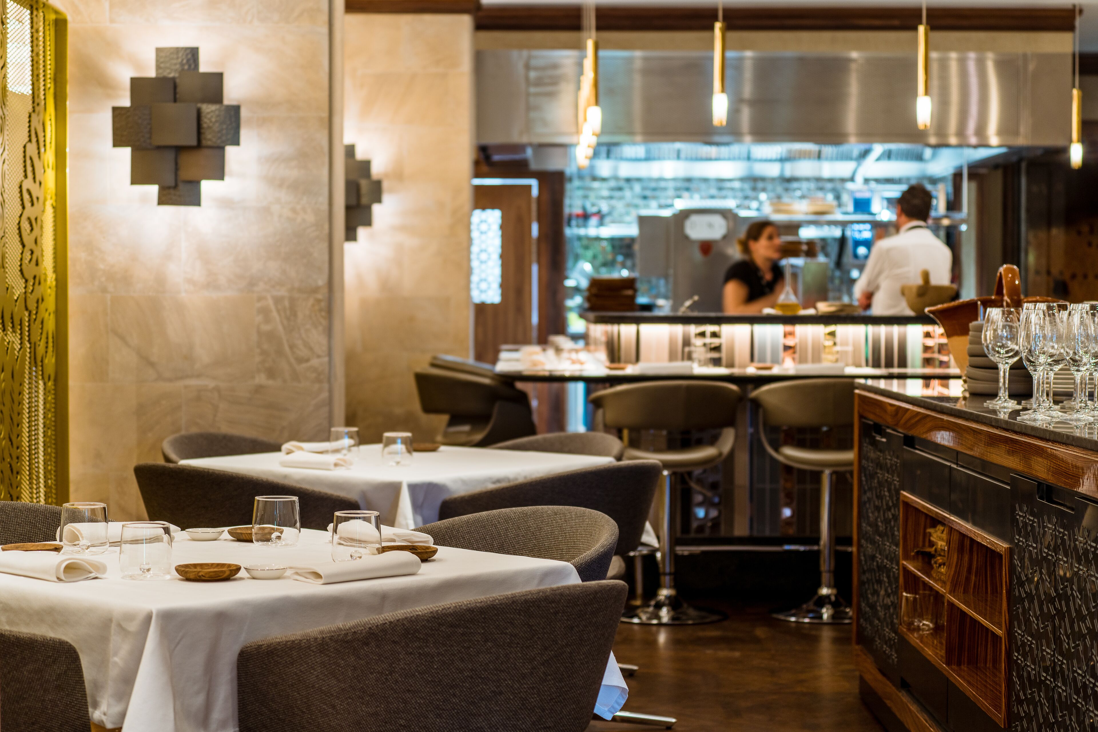 Un restaurante con mesas dispuestas para comer, una zona de bar con vasos y una cocina al fondo.