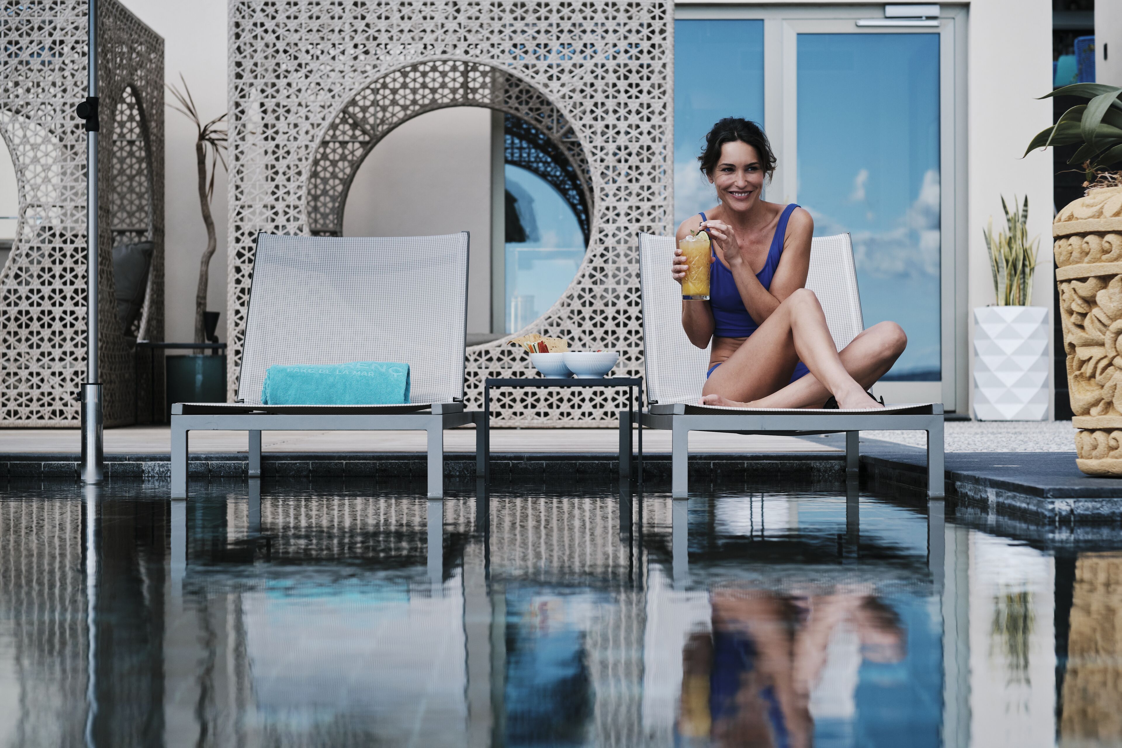 A poolside area with lounge chairs, a person holding a drink, and a decorative wall.