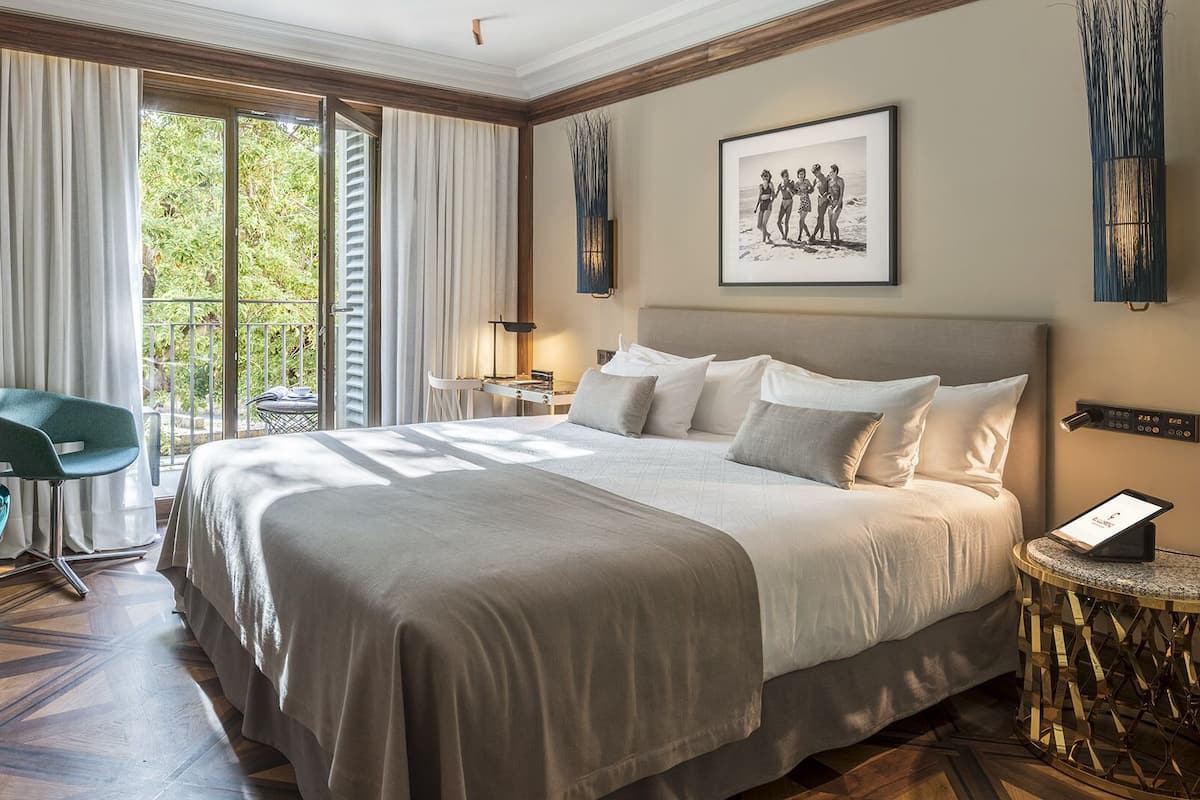 A hotel room with a large bed, a desk, a chair, and a view of greenery through the window.