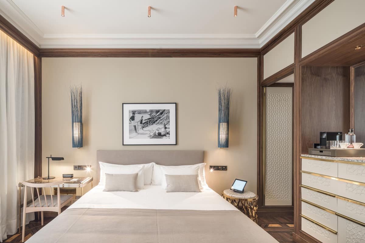 A hotel room with a large bed, a desk, a chair, a small round table, a laptop, and a nightstand with a lamp.