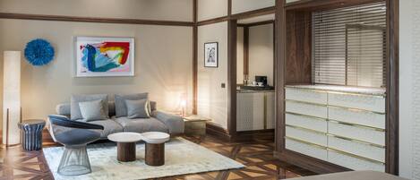 Una habitación de hotel moderna con sofá, una mesita de café, un objeto decorativo azul, un cuadro enmarcado, un armario con cajones y una ventana con persianas.