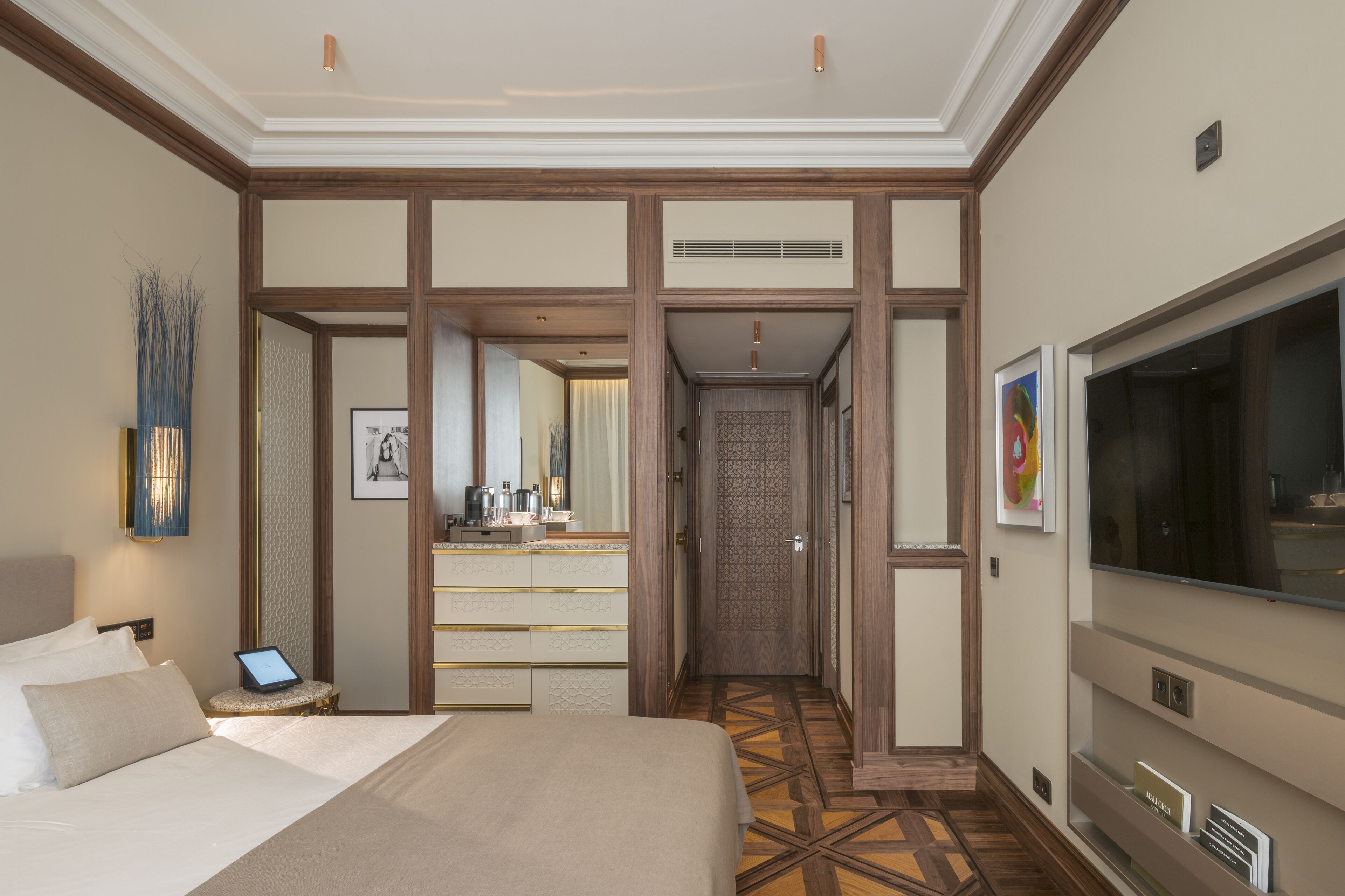 A hotel room with a large bed, a TV, a desk with a laptop, a mirror, and a hallway leading to a bathroom.