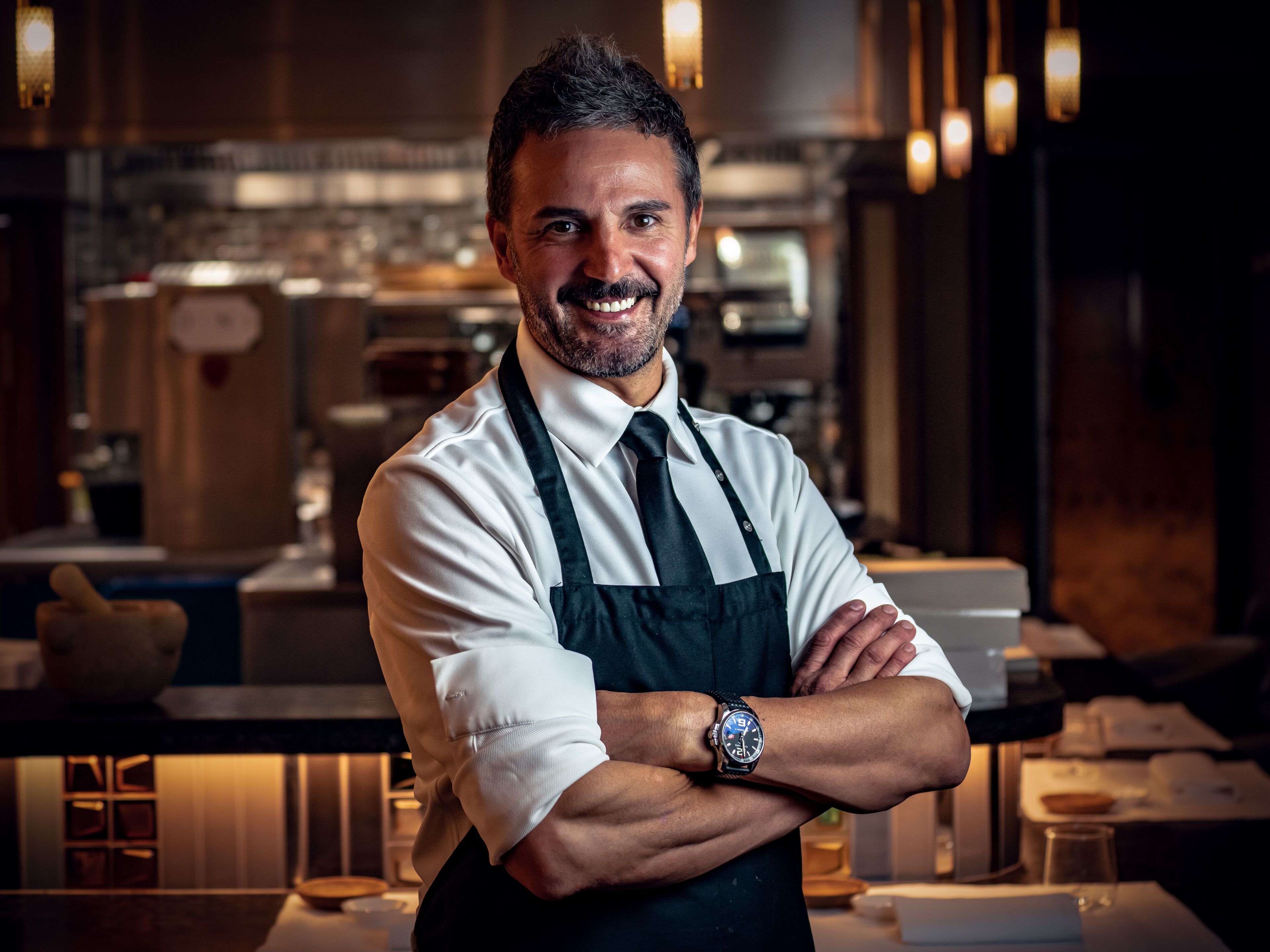 Una persona con camisa blanca y delantal negro, de brazos cruzados, en un ambiente interior cálidamente iluminado, posiblemente un restaurante o cafetería.