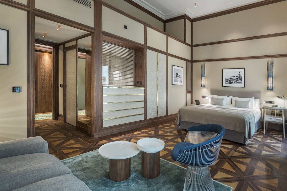 A hotel room with a bed, a chair, a small table, and a sofa. There are framed pictures on the walls and a window with blinds.