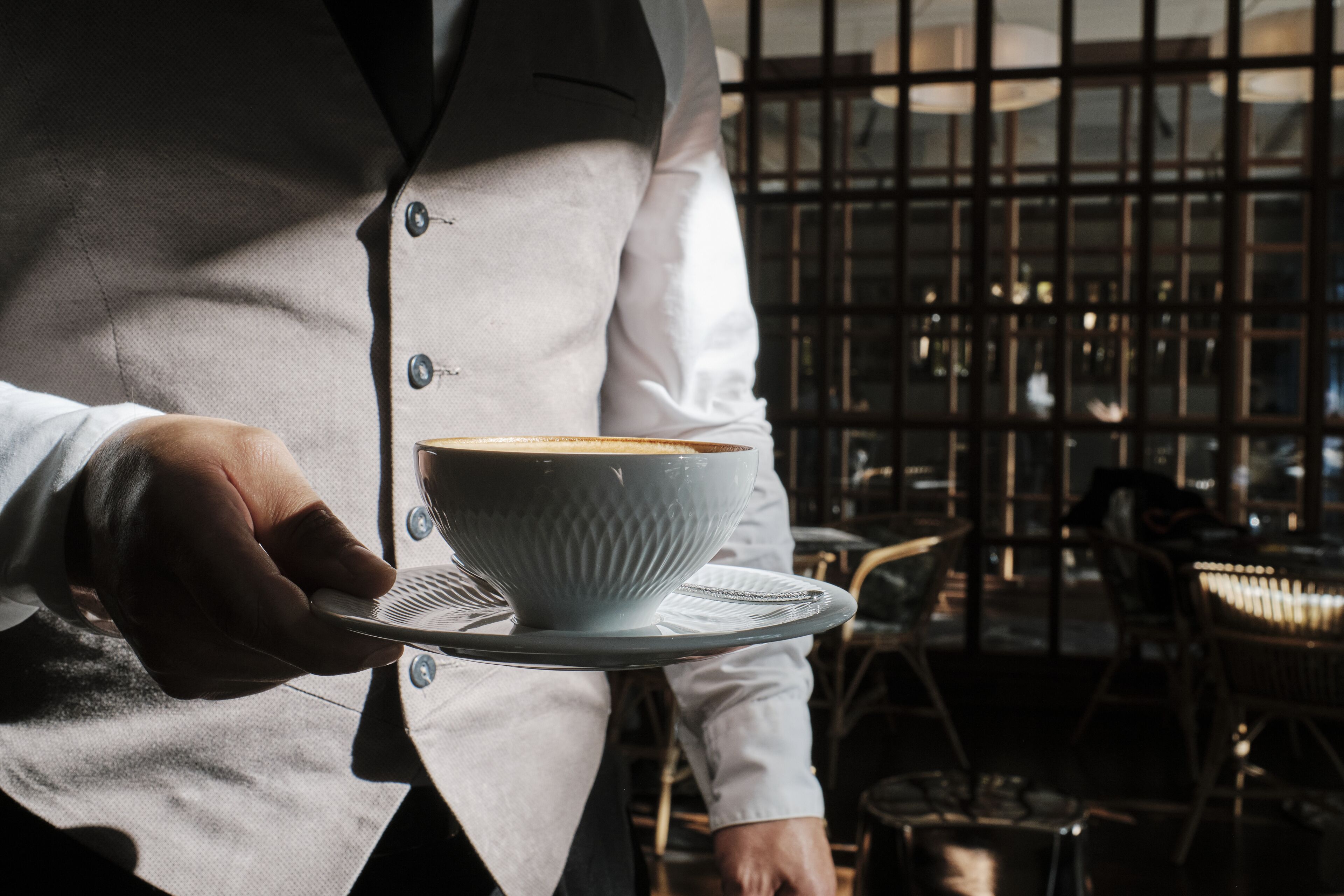 A person holding a white cup with a saucer, wearing a white shirt and a dark vest.