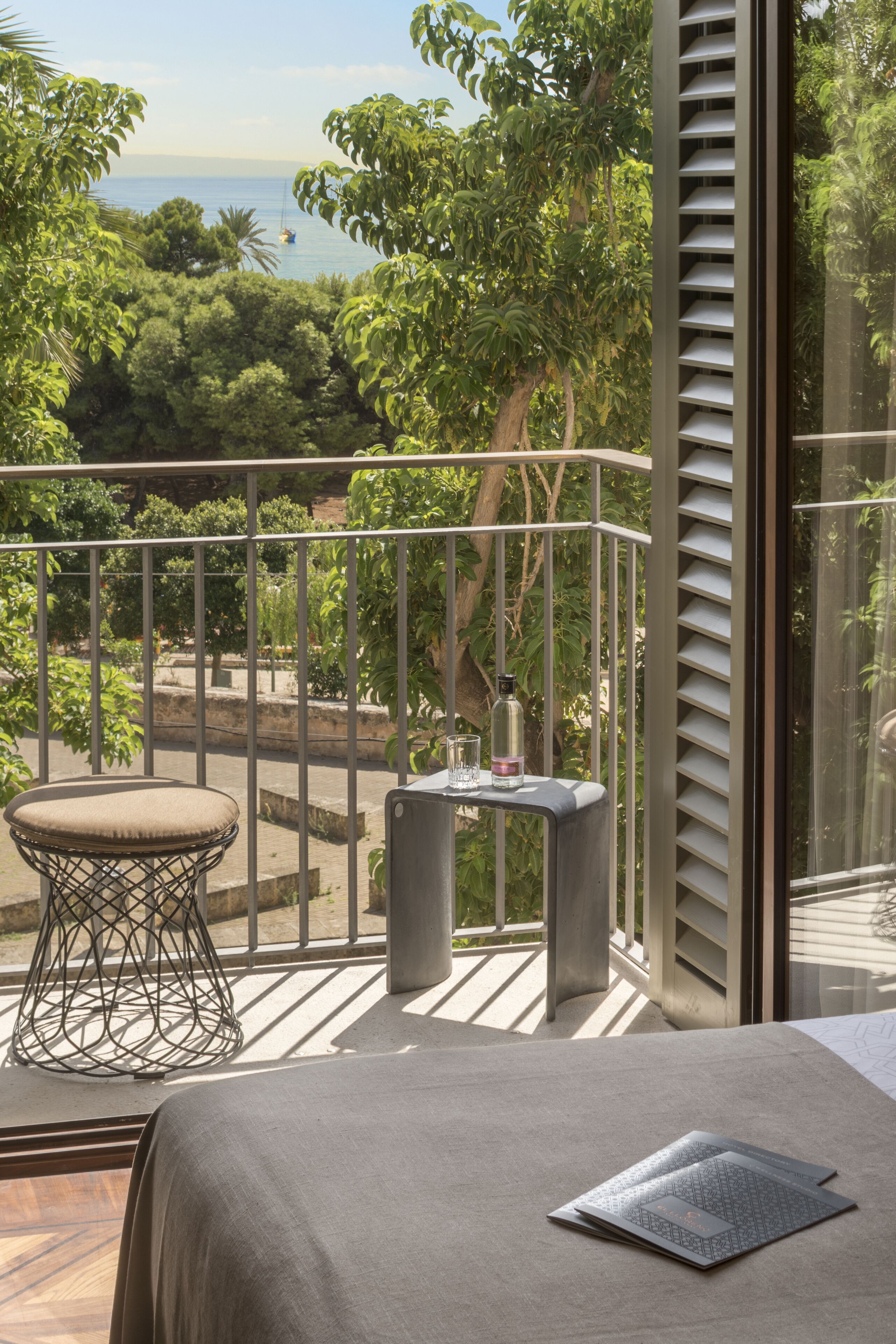 A balcony with a view of a tree-lined area and a distant boat. There is a cushioned chair and a small table with a bottle and glass.