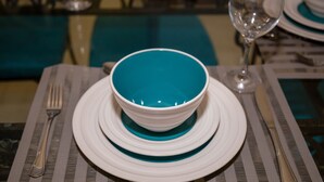 Une table dressée avec un bol turquoise, des assiettes blanches, une fourchette et un verre à vin.