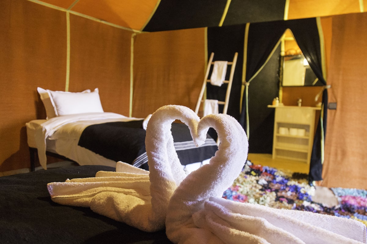 A bed with a heart-shaped towel arrangement, a wooden headboard, and a small bedside table with a lamp.