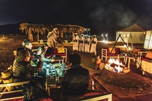 Un grupo de personas cenando al aire libre de noche, con una hoguera y tiendas de campaña al fondo.