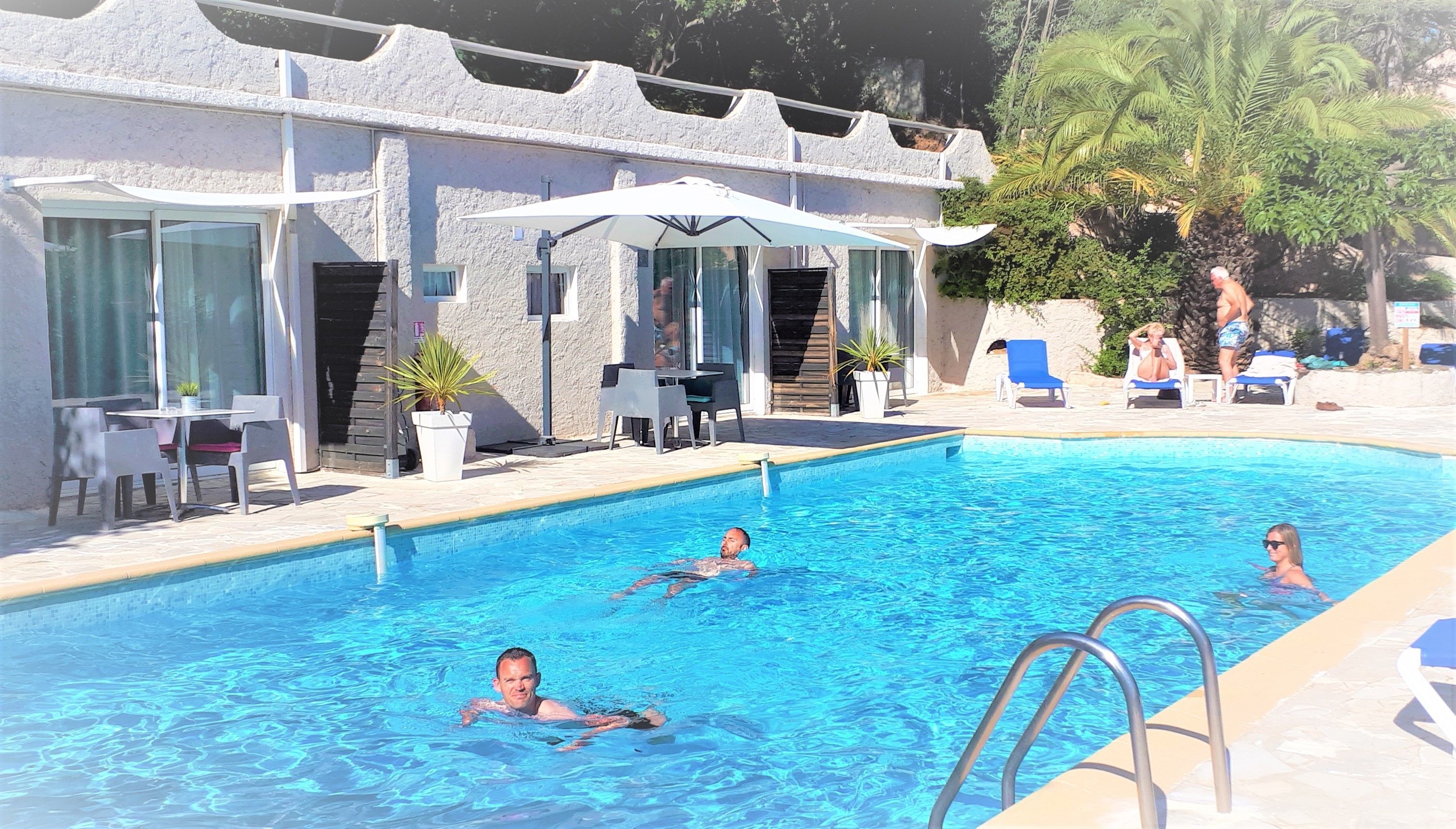 En swimmingpool med solstolar och parasoller, omgiven av en byggnad med stora fönster och utomhusmöbler.