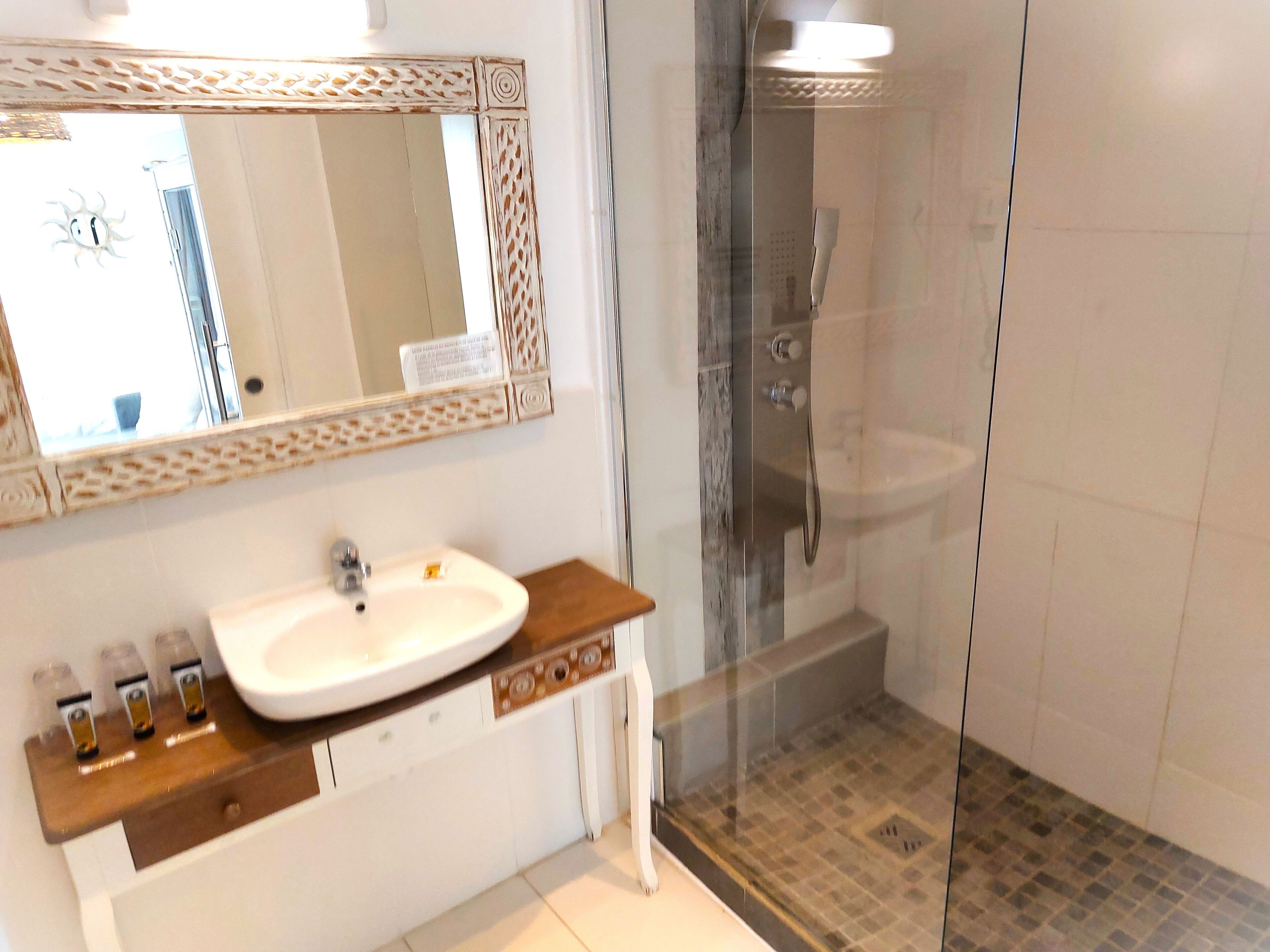 Une salle de bain avec un lavabo blanc, un miroir doté d’un cadre décoratif et un espace douche séparé par une porte en verre.