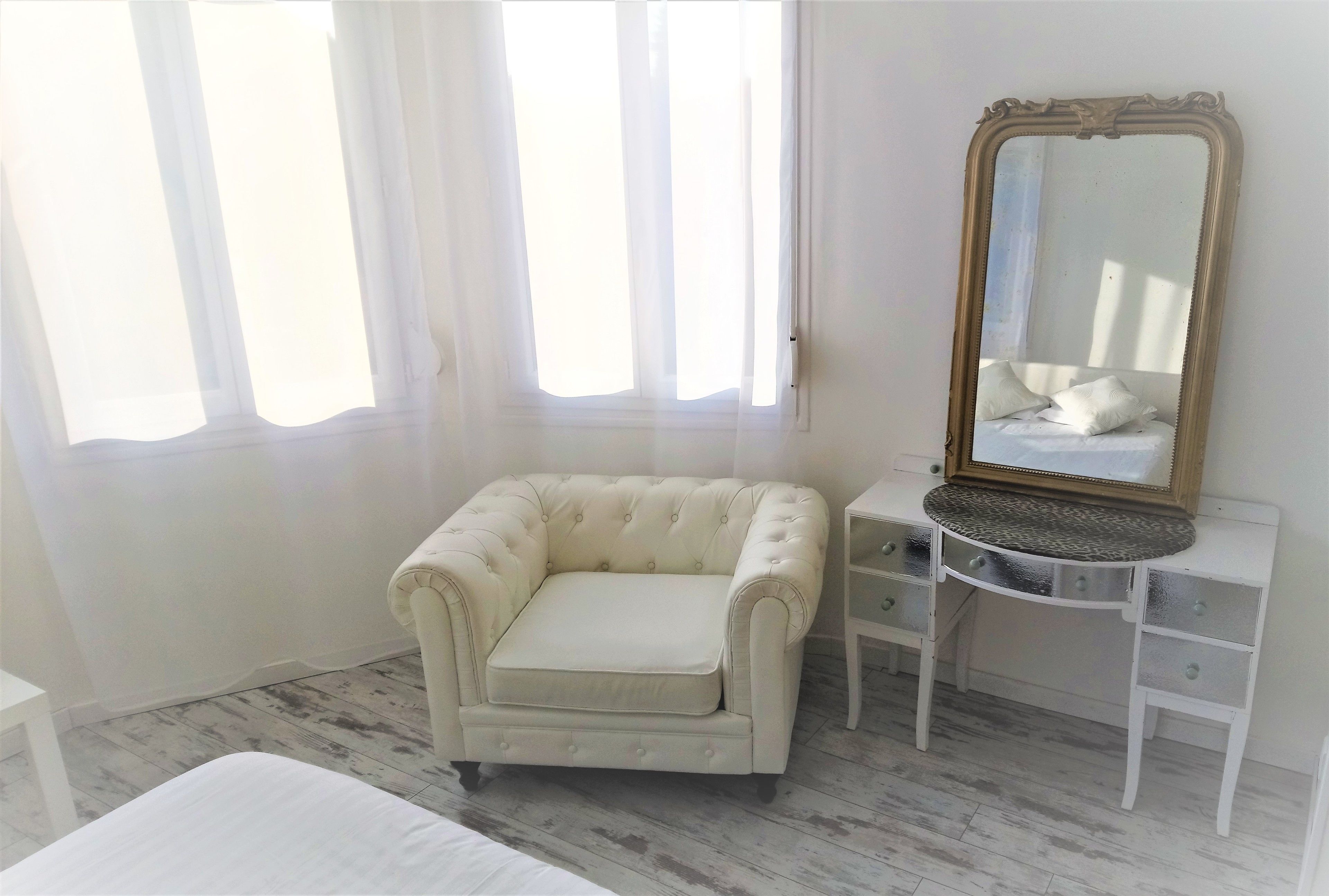 Une pièce comprenant un fauteuil blanc, une coiffeuse avec un miroir et un lit.