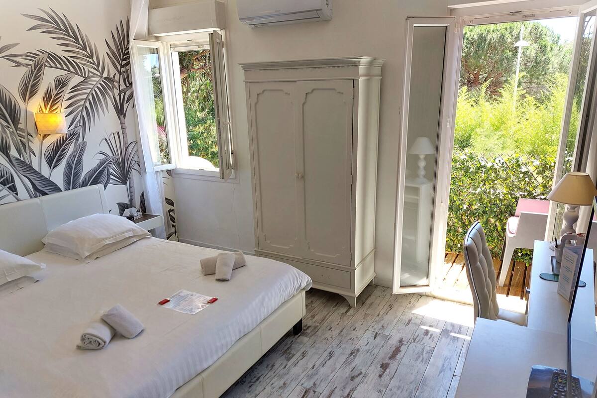Un dormitorio con cama, armario, escritorio con lámpara y vistas a la vegetación a través de una puerta abierta.