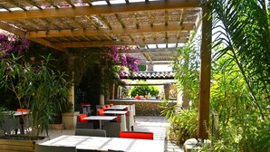 Un patio al aire libre con mesas y sillas, rodeado de plantas y una pérgola de madera.