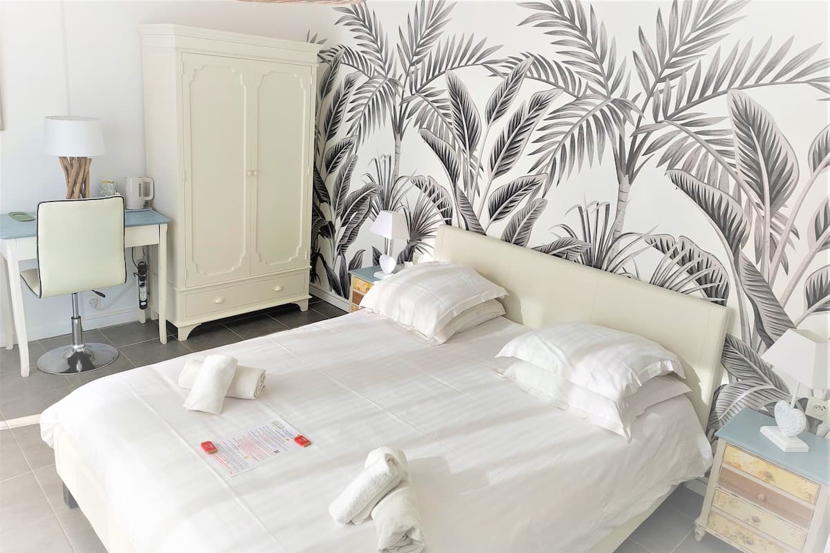 Una cama bien hecha con ropa y toallas blancas, un armario blanco, un escritorio con lámpara y una pared con estampado de hojas tropicales.