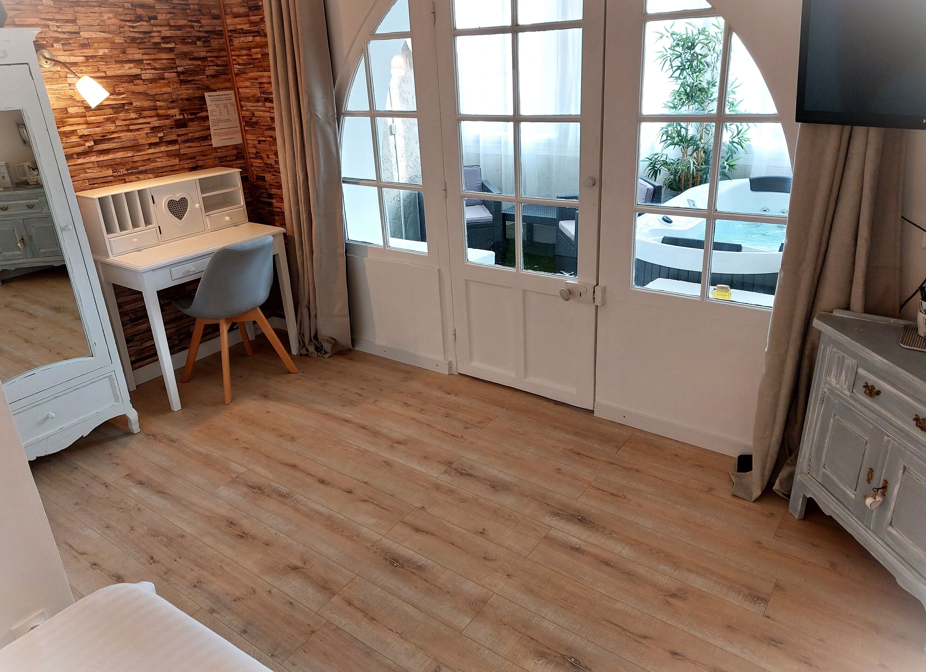 Une pièce avec un plancher en bois, un bureau avec une chaise, une commode blanche et une grande fenêtre en arc surmontée de rideaux.