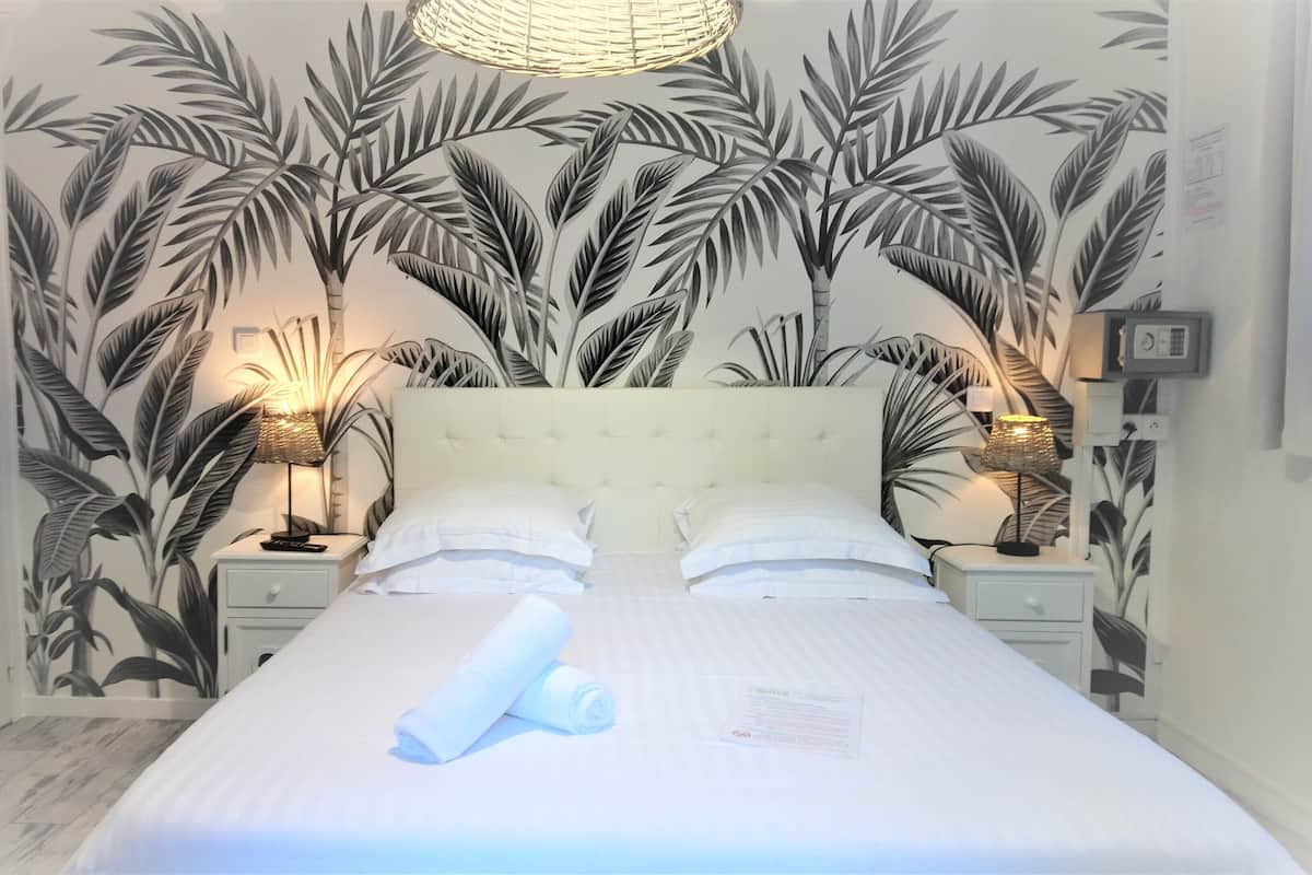 Una cama bien tendida con sábanas blancas, dos mesitas de noche con lámparas, y una pared con diseño de hojas tropicales.