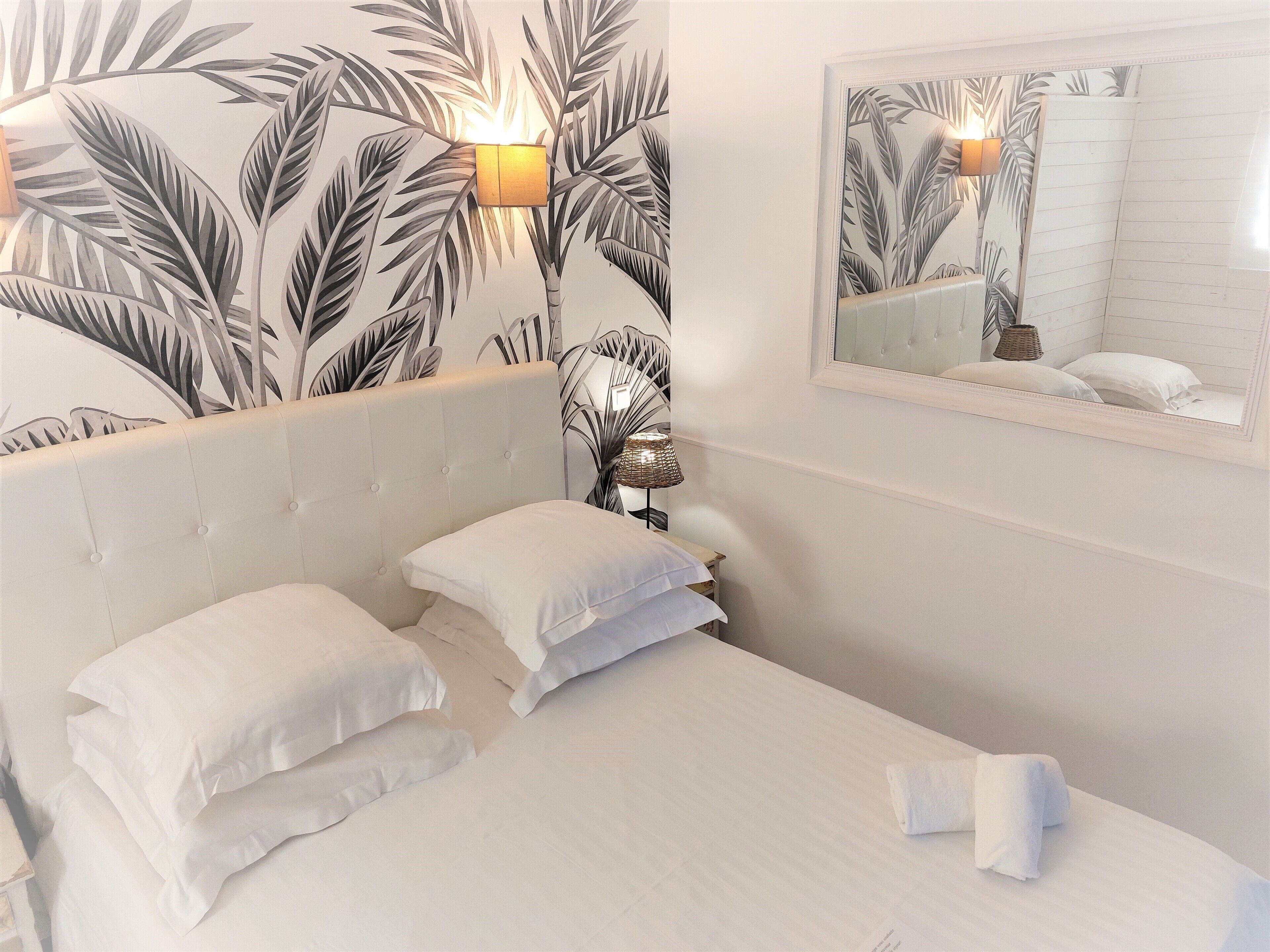 A hotel room with a bed, two pillows, a nightstand with a lamp, a mirror, and a wall with a tropical leaf pattern.