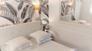 A hotel room with a bed, two pillows, a nightstand with a lamp, a mirror, and a wall with a tropical leaf pattern.