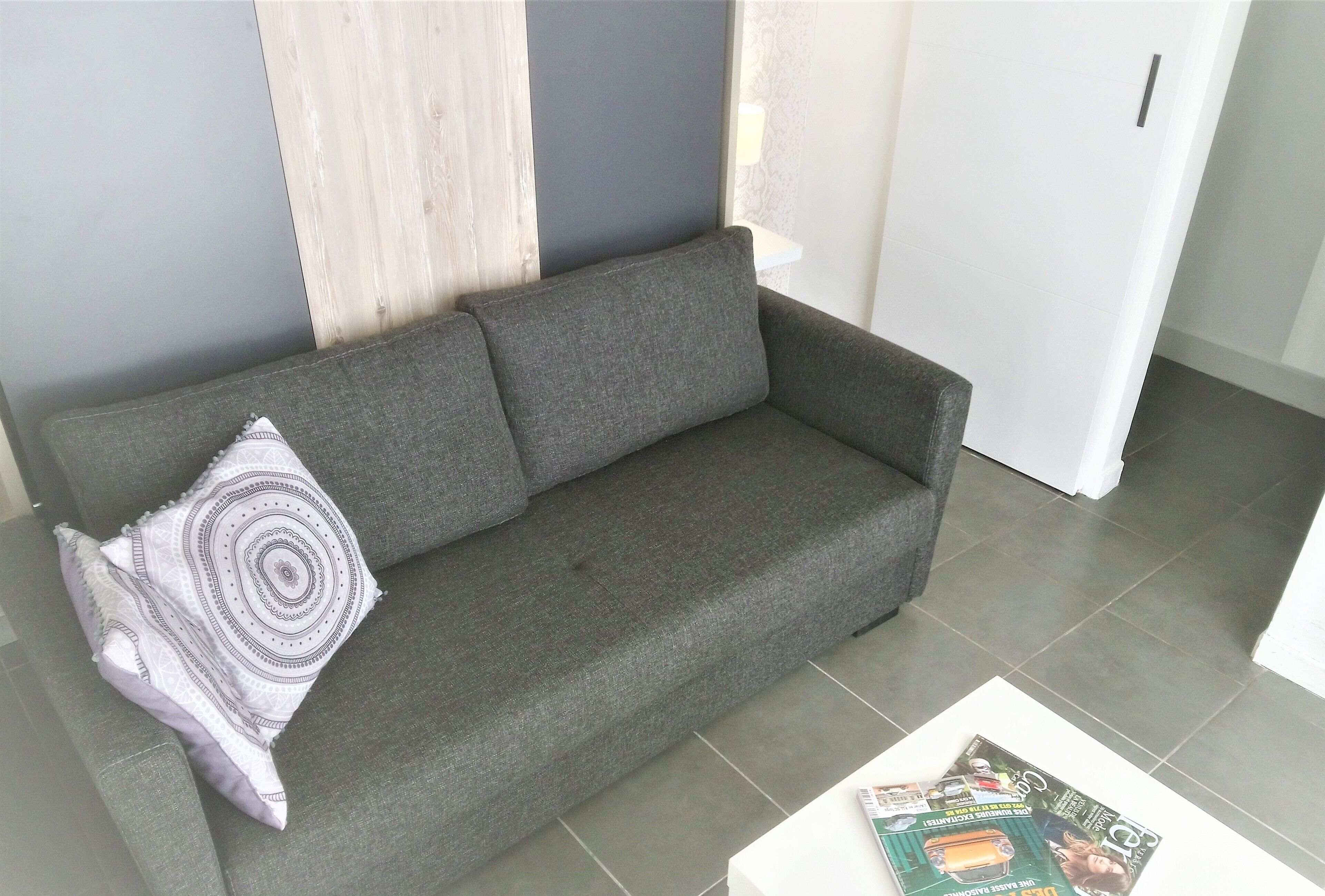 Ein graues Sofa mit einem gemusterten Kissen, ein weißer Couchtisch mit einer Zeitschrift und eine weiße Tür in einem Raum mit grauen Wänden und Fliesenboden.