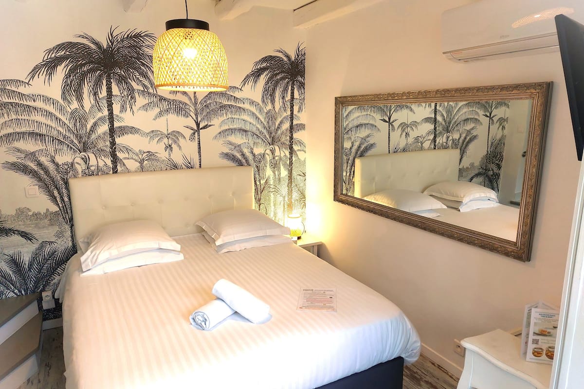 Una cama bien hecha con sábanas blancas y una toalla enrollada, una lámpara colgante, un espejo grande y una pared con un mural tropical.