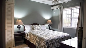 Un dormitorio con cama, dos mesitas de noche con lámparas, ventilador de techo, equipo de aire acondicionado, ventana con cortinas y puerta con contraventanas.