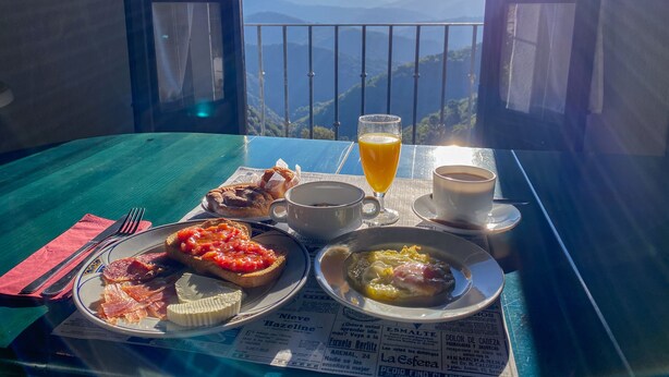 Ett bord med frukostmat, ett glas apelsinjuice och en kopp kaffe, med en vacker utsikt över bergen från balkongen.