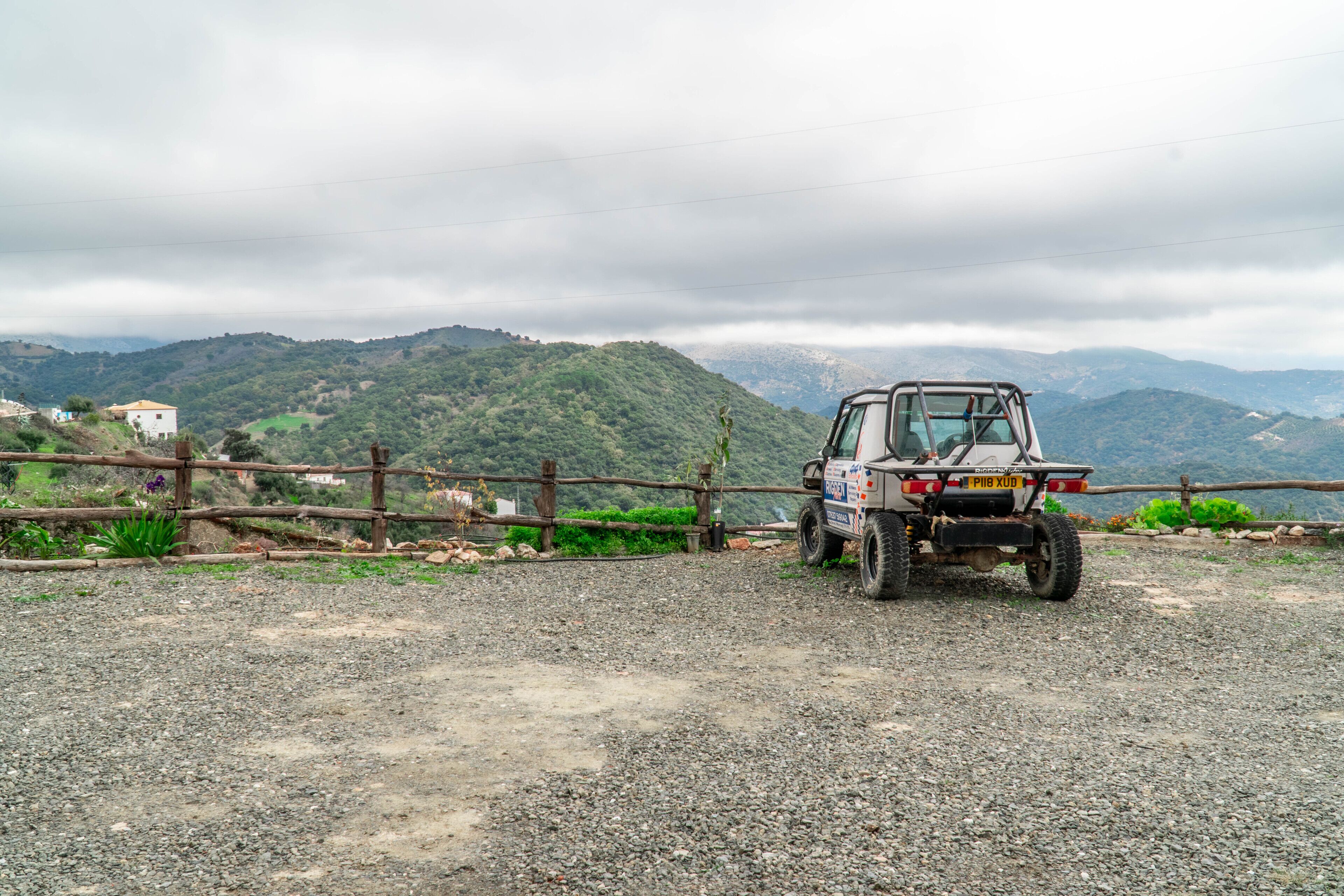 A rugged off-road vehicle parked on a gravel path with a wooden fence and a mountainous backdrop.