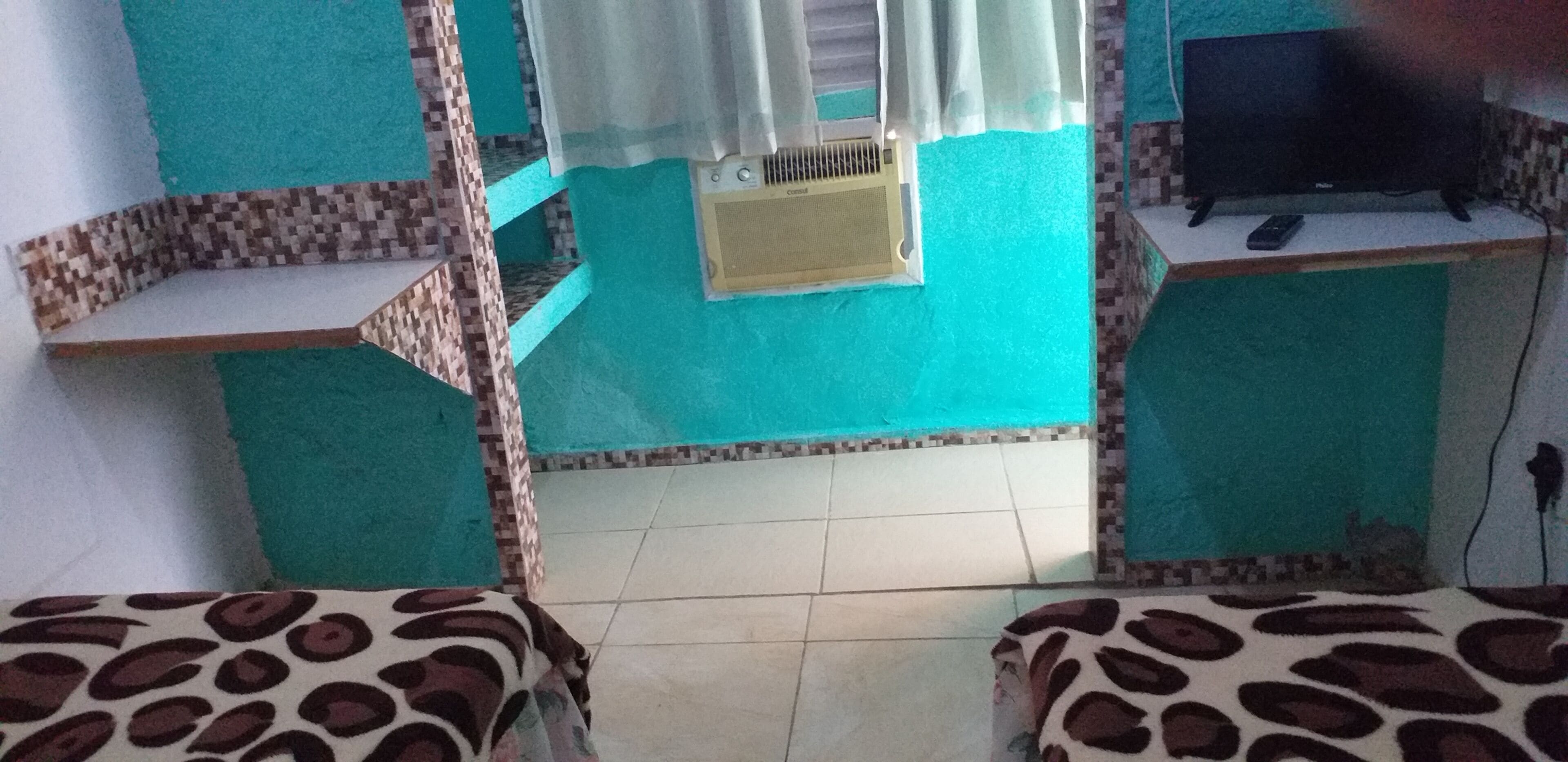 Una habitación con paredes verde azulado, piso de baldosas, una ventana con cortina blanca, un pequeño equipo de aire acondicionado, un televisor sobre un mueble y dos camas con estampado de animales.
