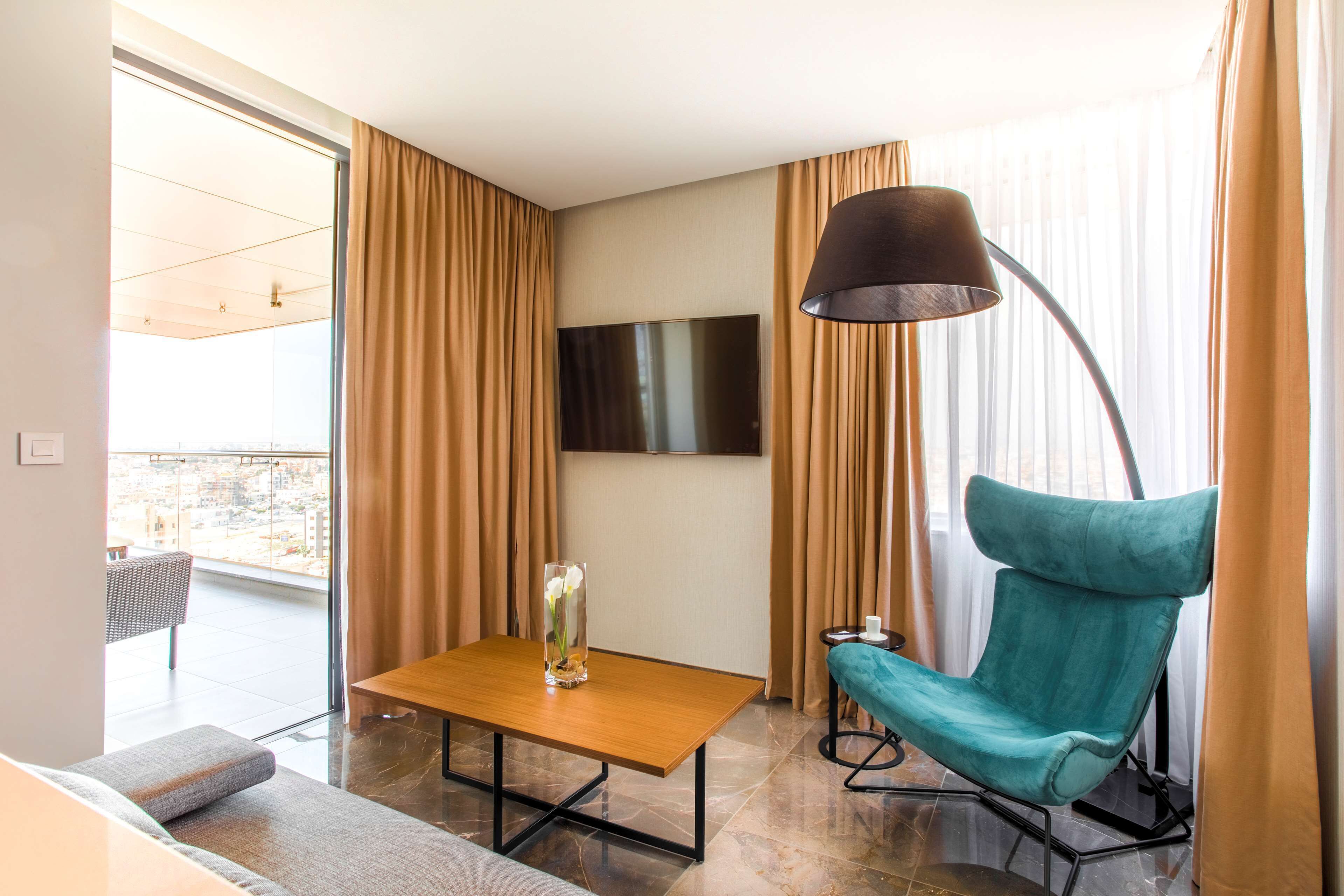 Una habitación de hotel moderna con un ventanal grande, un televisor de pantalla plana, un sillón verde azulado, una mesa de madera y una lámpara de pie.