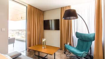 Una habitación de hotel moderna con un ventanal grande, un televisor de pantalla plana, un sillón verde azulado, una mesa de madera y una lámpara de pie.