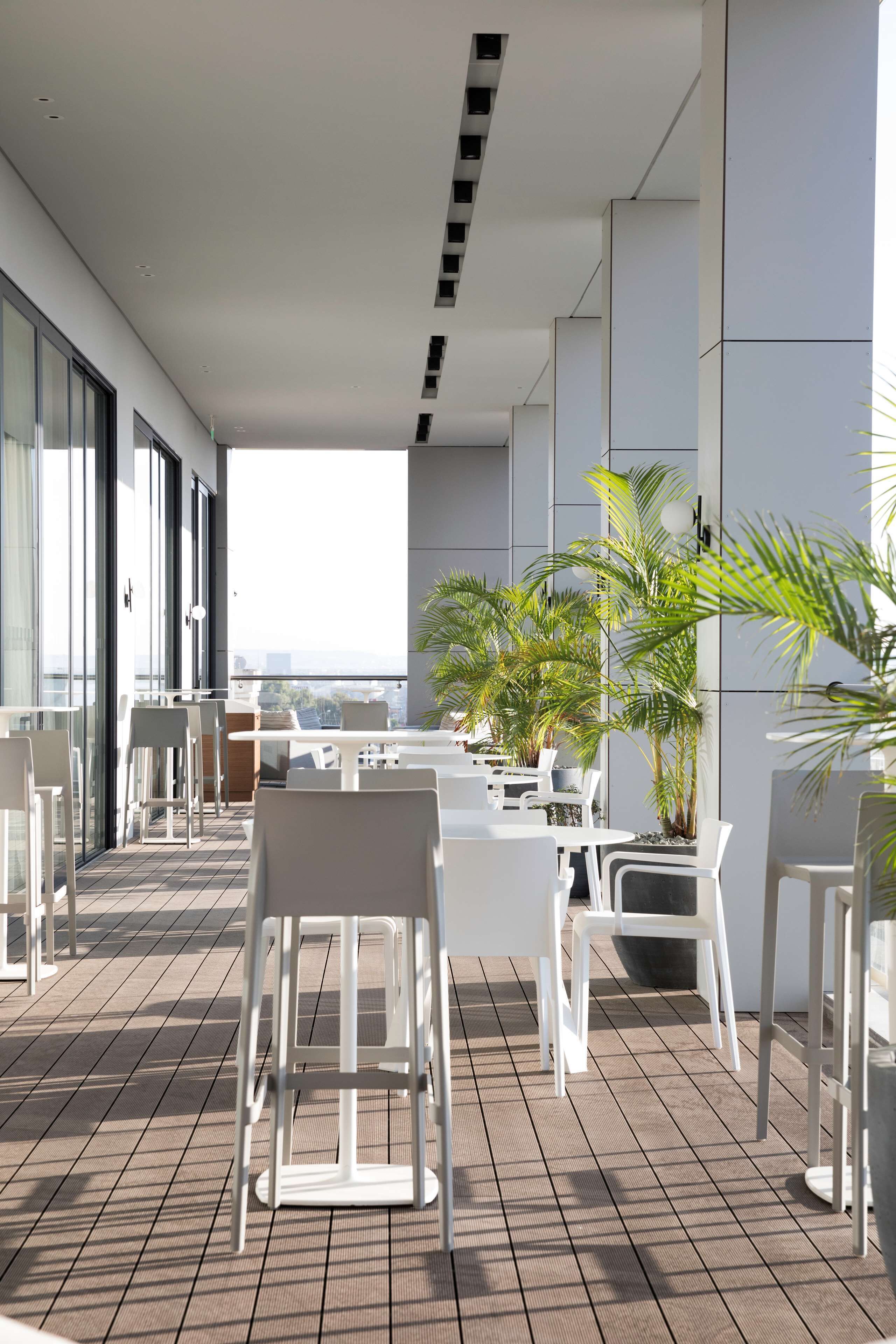 A modern outdoor patio with white furniture, potted plants, and large windows offering a city view.