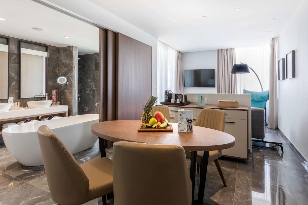 A modern hotel room with a round dining table, a large bathtub, and a kitchen area with a flat-screen TV.