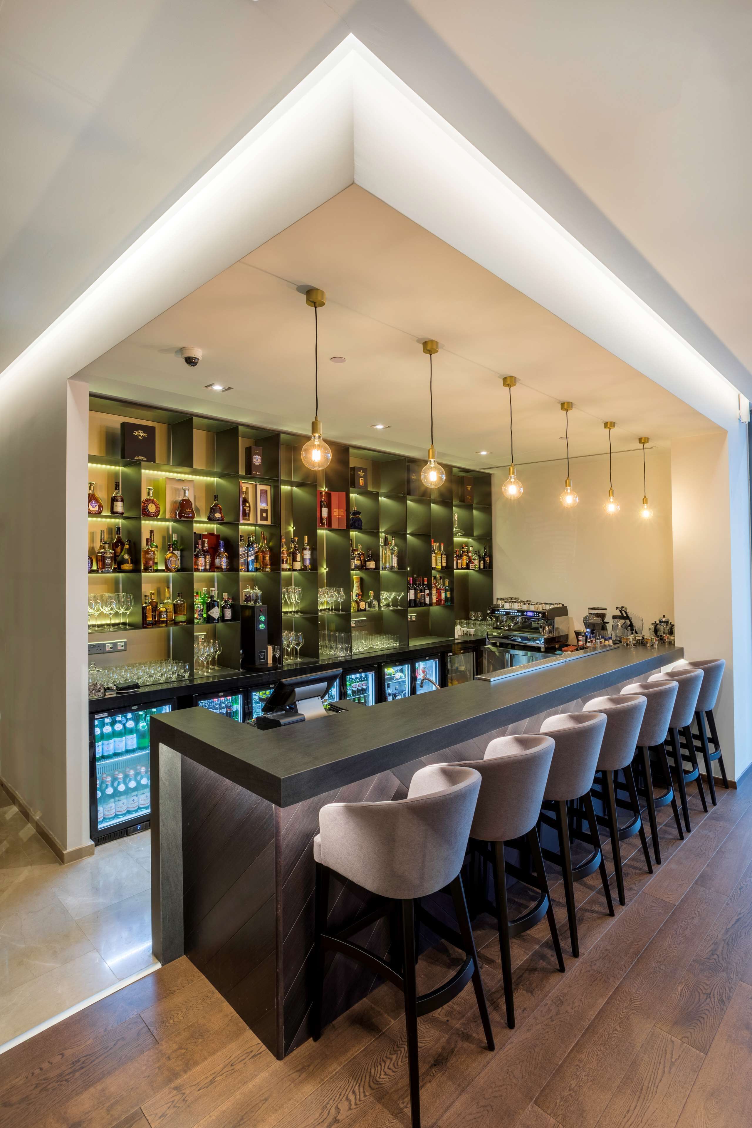 Un espace bar moderne doté d’un long comptoir, de nombreux tabourets et d’une étagère bien garnie de bouteilles et de verres de toutes sortes.