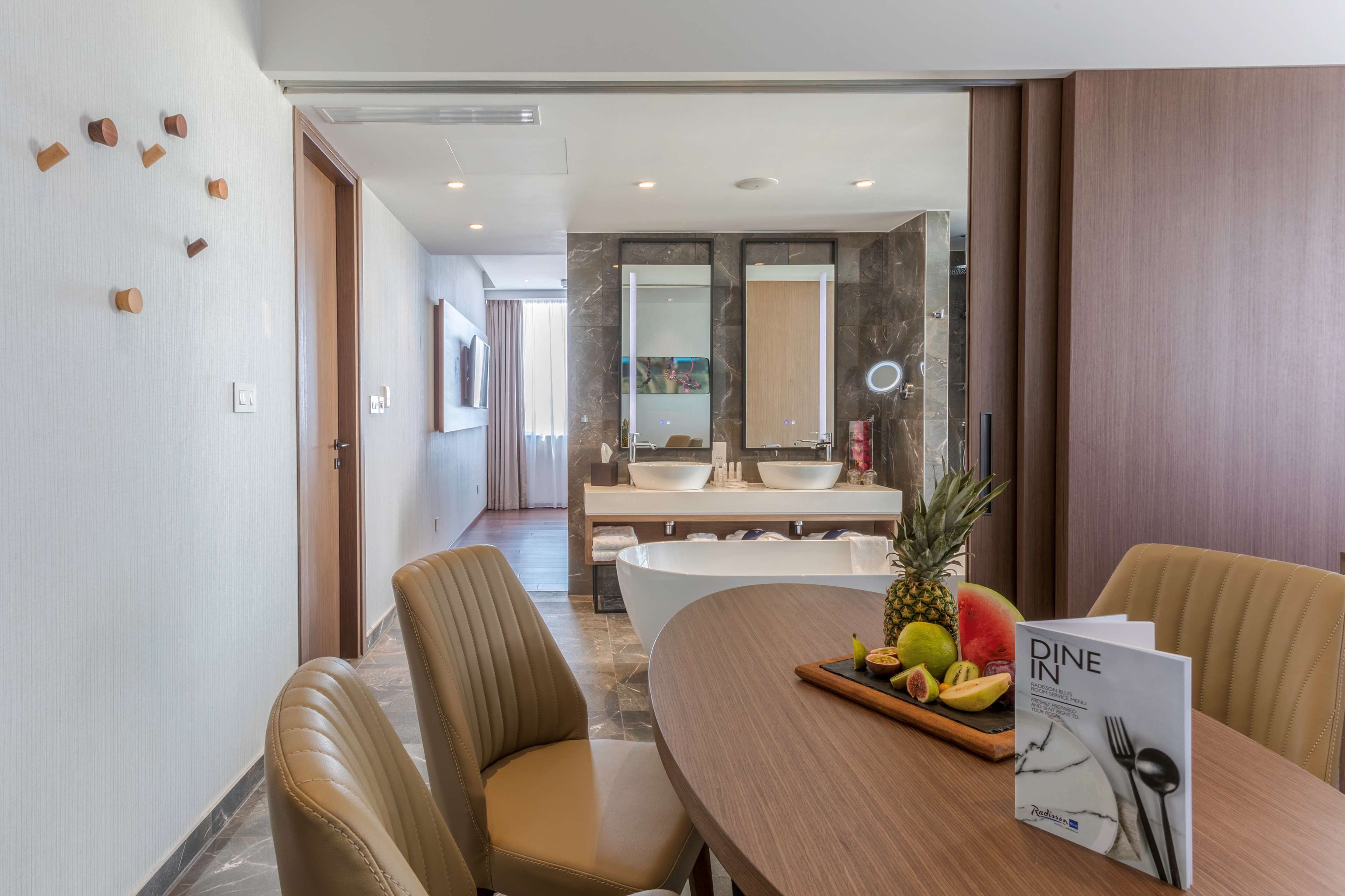 A modern hotel room with a dining table set for two, a fruit tray, and a 'Dine In' menu.