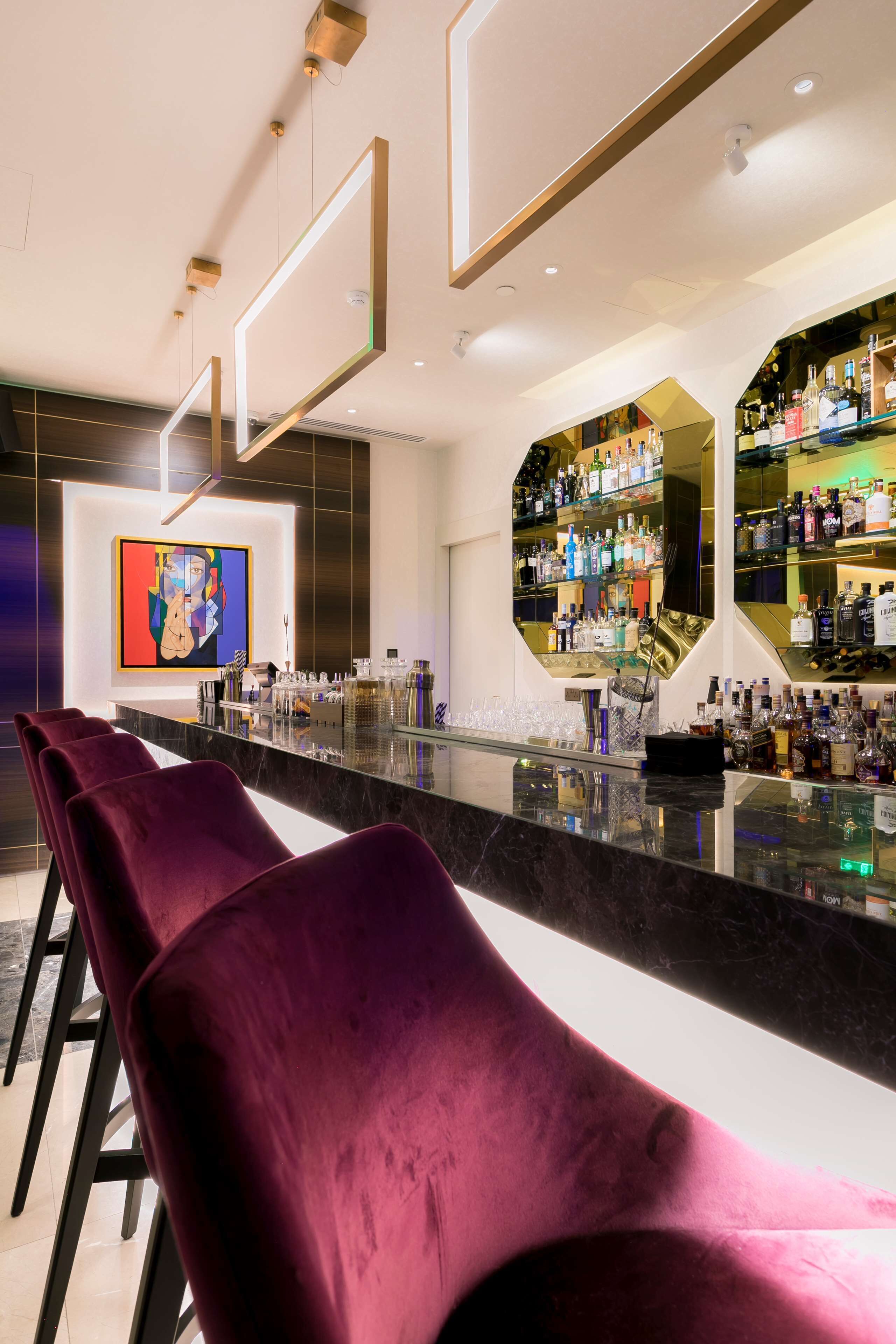 A bar area with a long counter, multiple shelves of bottles, and a large painting on the wall.