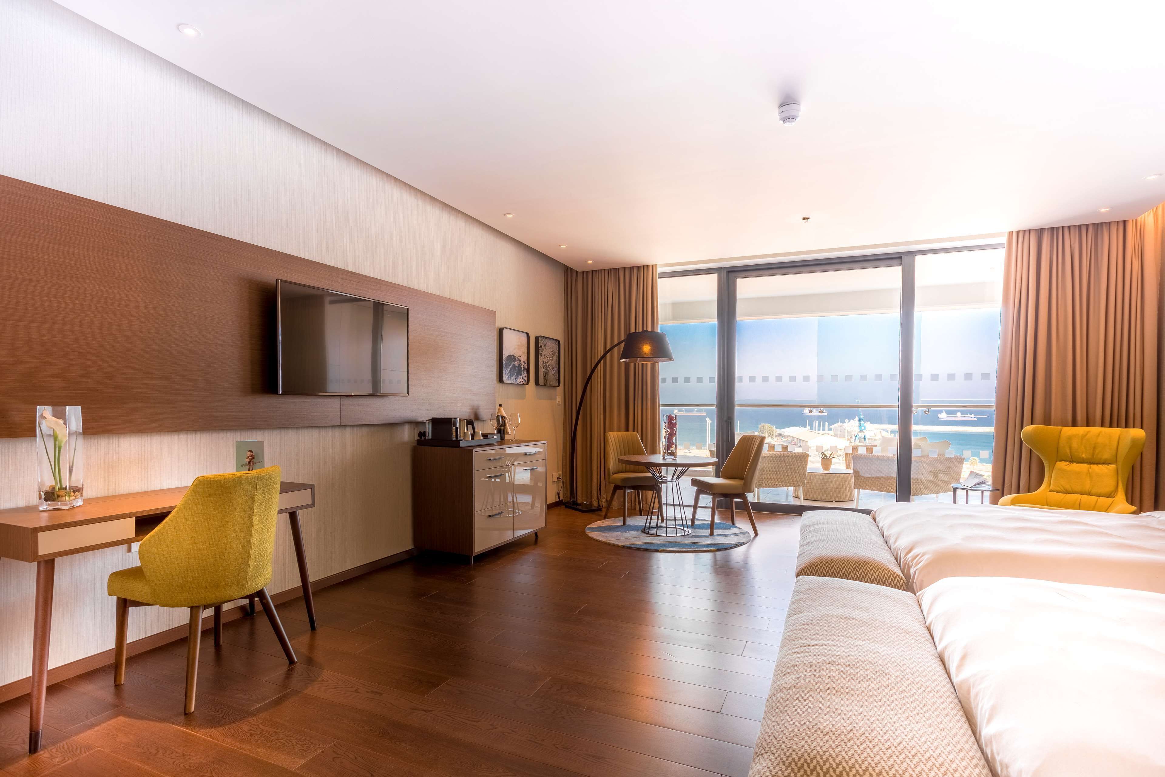 A hotel room with a large bed, a desk with a chair, a TV, a dining area with chairs, and a view of the city.