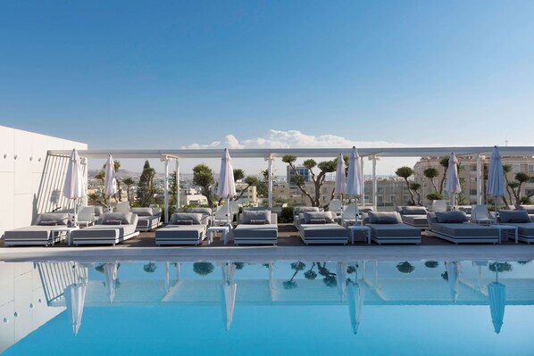 A rooftop pool area with sun loungers and umbrellas, clear blue sky, and a cityscape in the background.